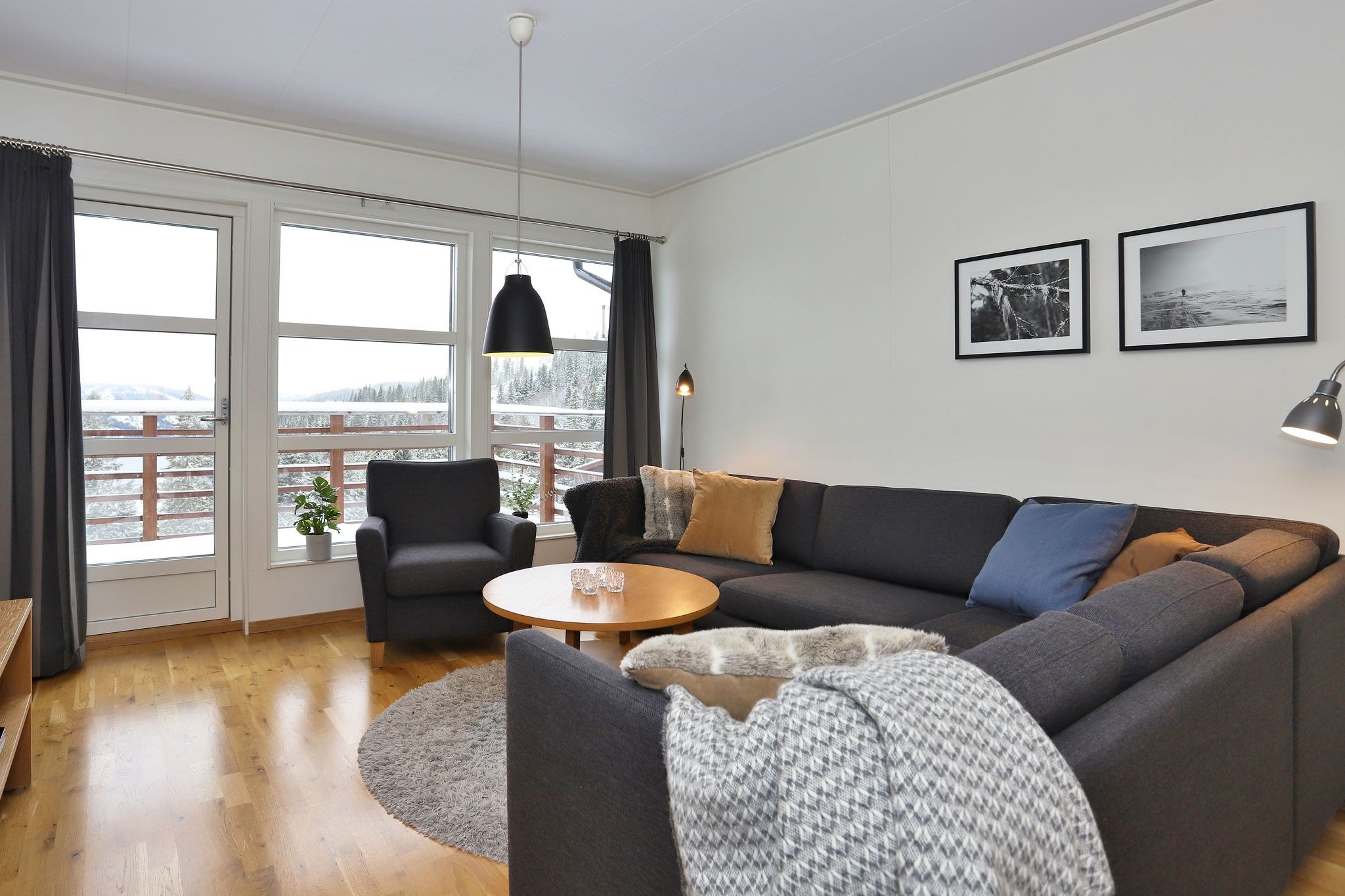 En moderne stue med en stor grå sofa, et rundt træbord og hyggelige tæpper. Store vinduer lukker naturligt lys ind og tilbyder en smuk udsigt over skoven. To sort-hvide fotos hænger på den hvide væg.