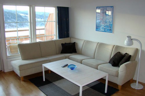 En lys stue med en beige hjørnesofa, to brune hynder, et hvidt sofabord med en blå skål og fjernbetjening, en gulvlampe, vægkunst og store vinduer, der viser en snedækket udendørs balkon.