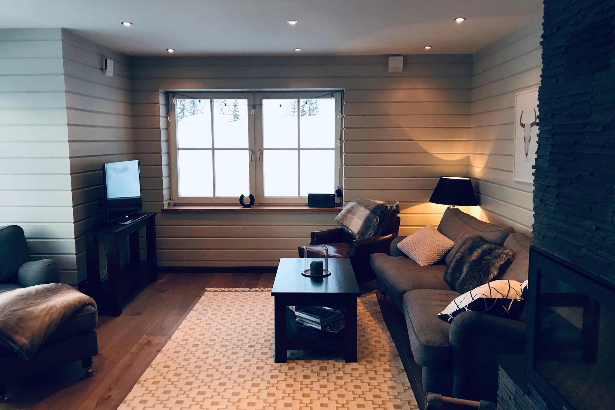 Hyggelig stue med en beige sofa, lænestole, et mørkt sofabord, TV på et bord og et mønstret tæppe. Et stort vindue lukker lys ind, og en lampe, dekoration og pejs tilføjer varme til rummet.