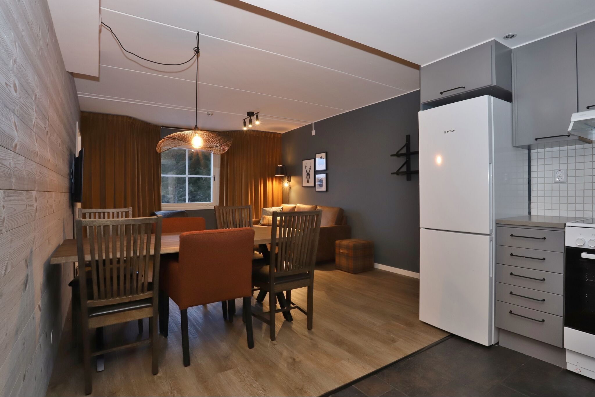 Moderne åben spise- og opholdsstue med et træbord, seks forskellige stole, en brun sofa, et stort vindue med gardiner og et køkkenområde med et hvidt køleskab og komfur.