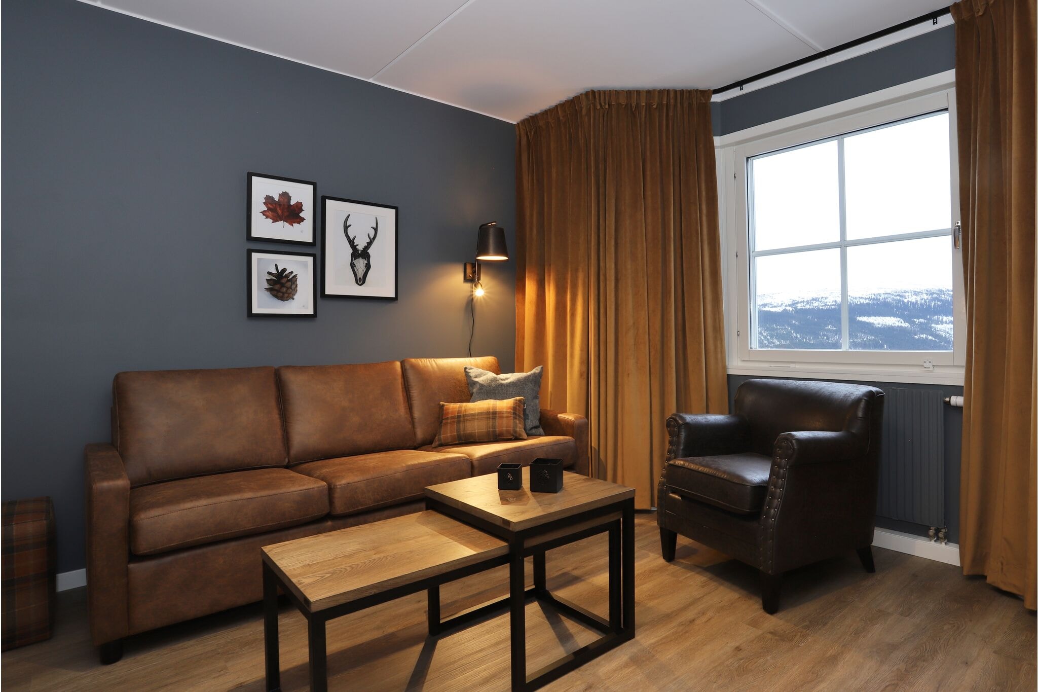 En hyggelig stue med en brun lædersofa, en mørk lænestol, sofaborde i træ, indrammet kunst på en grå væg, gyldne gardiner og et vindue med udsigt over en sneklædt bjergudsigt.
