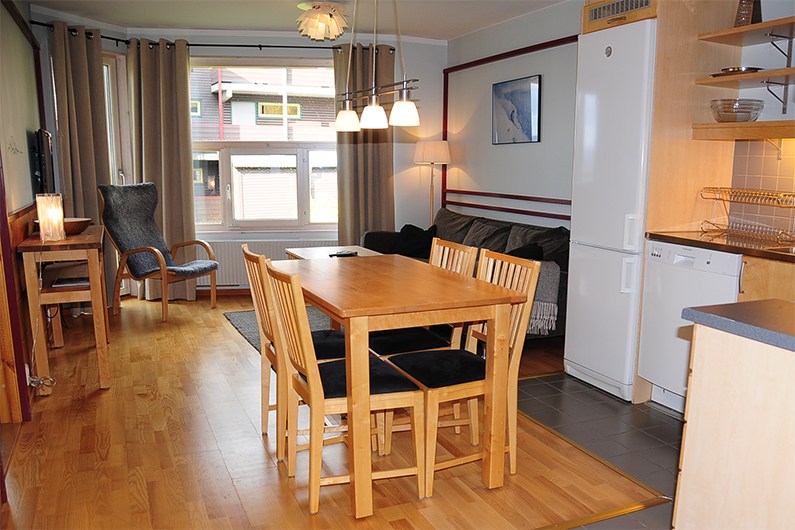 En hyggelig spise- og opholdsstue med et træbord og fire stole, en sofa, lænestol og et stort vindue med beige gardiner. Køkkenområdet er synligt med køleskab, opvaskemaskine og åbne hylder.