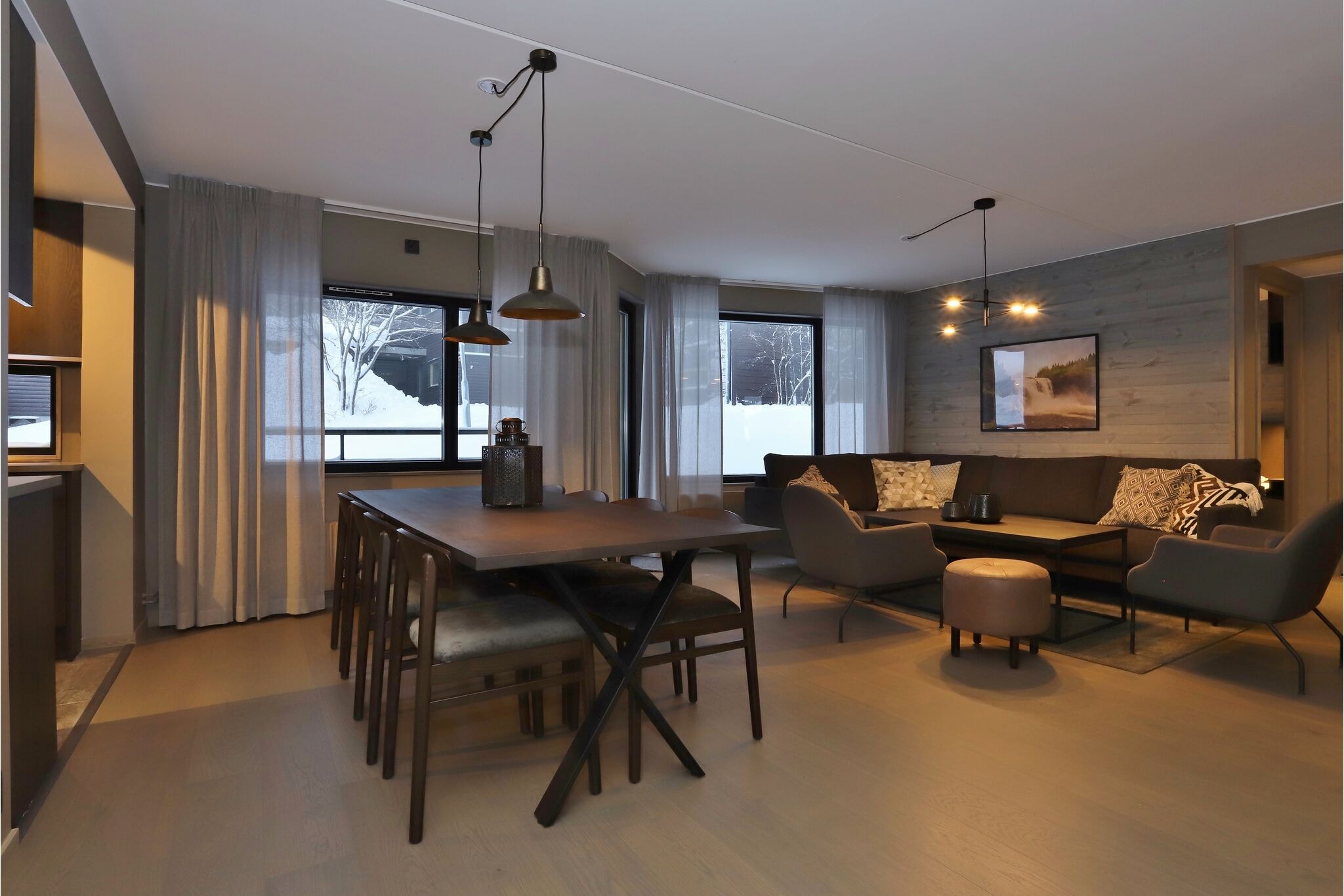 Moderne opholds- og spisestue med trægulve, et mørkt spisebord med stole, en grå hjørnesofa, lænestole, en rund puf og store vinduer med sne synlig udenfor. Varm, stemningsfuld belysning overalt.
