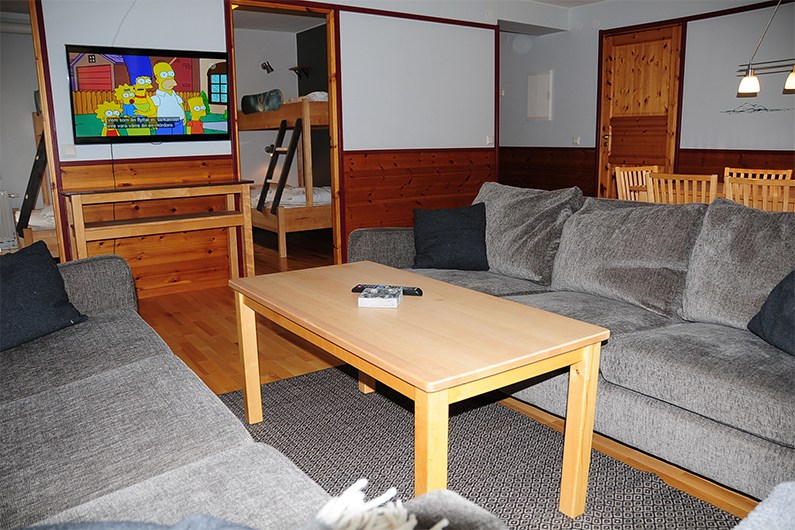En hyggelig stue med grå sofaer omkring et træbord, et tv med The Simpsons, køjesenge i baggrunden, trævægge og en spiseplads med stole og et bord.