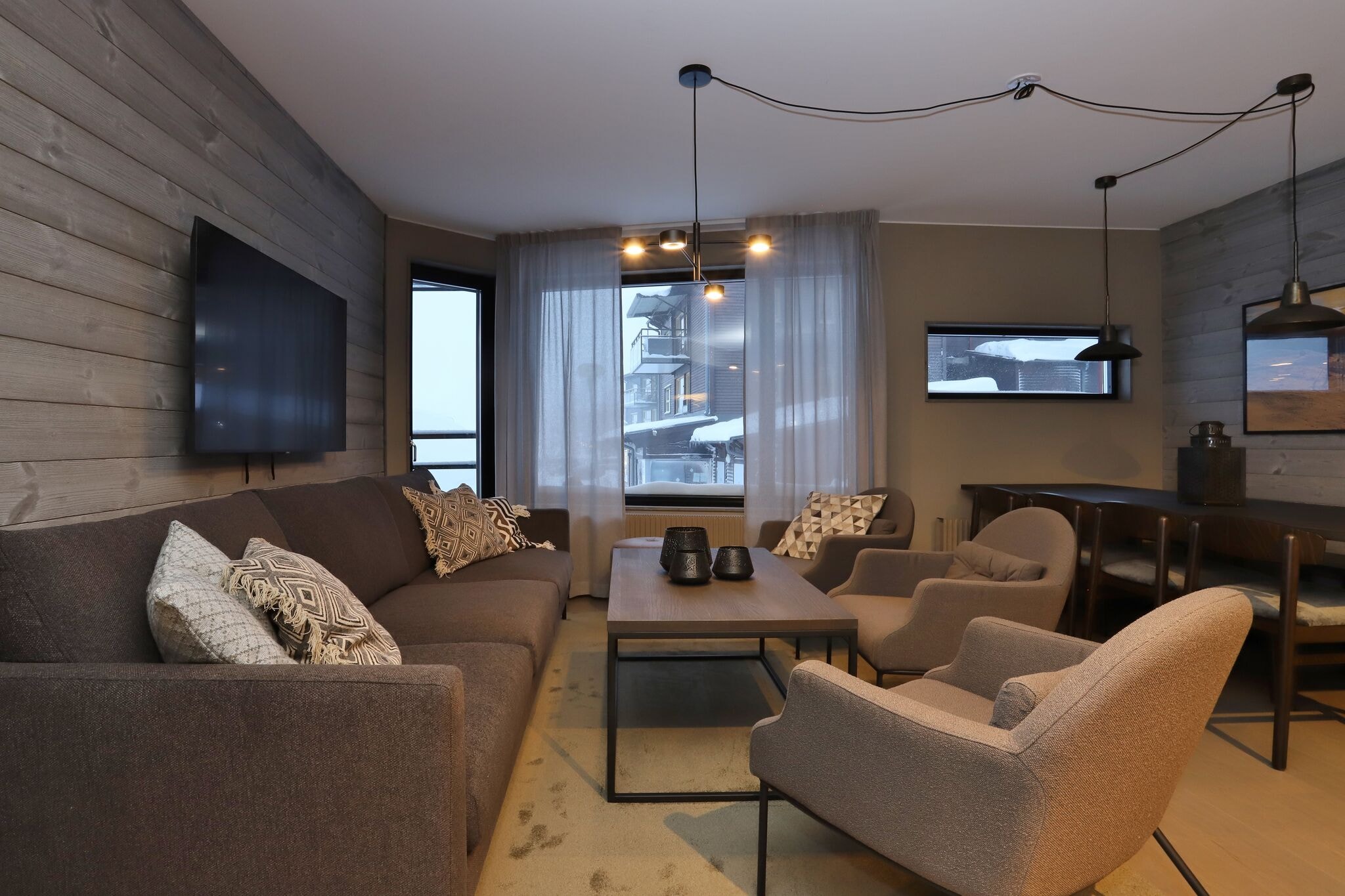 Moderne stue med en grå sofa, to lænestole, et sofabord i træ, vægmonteret TV og store vinduer med tynde gardiner, der lukker naturligt lys ind og viser et snedækket udendørssceneri.