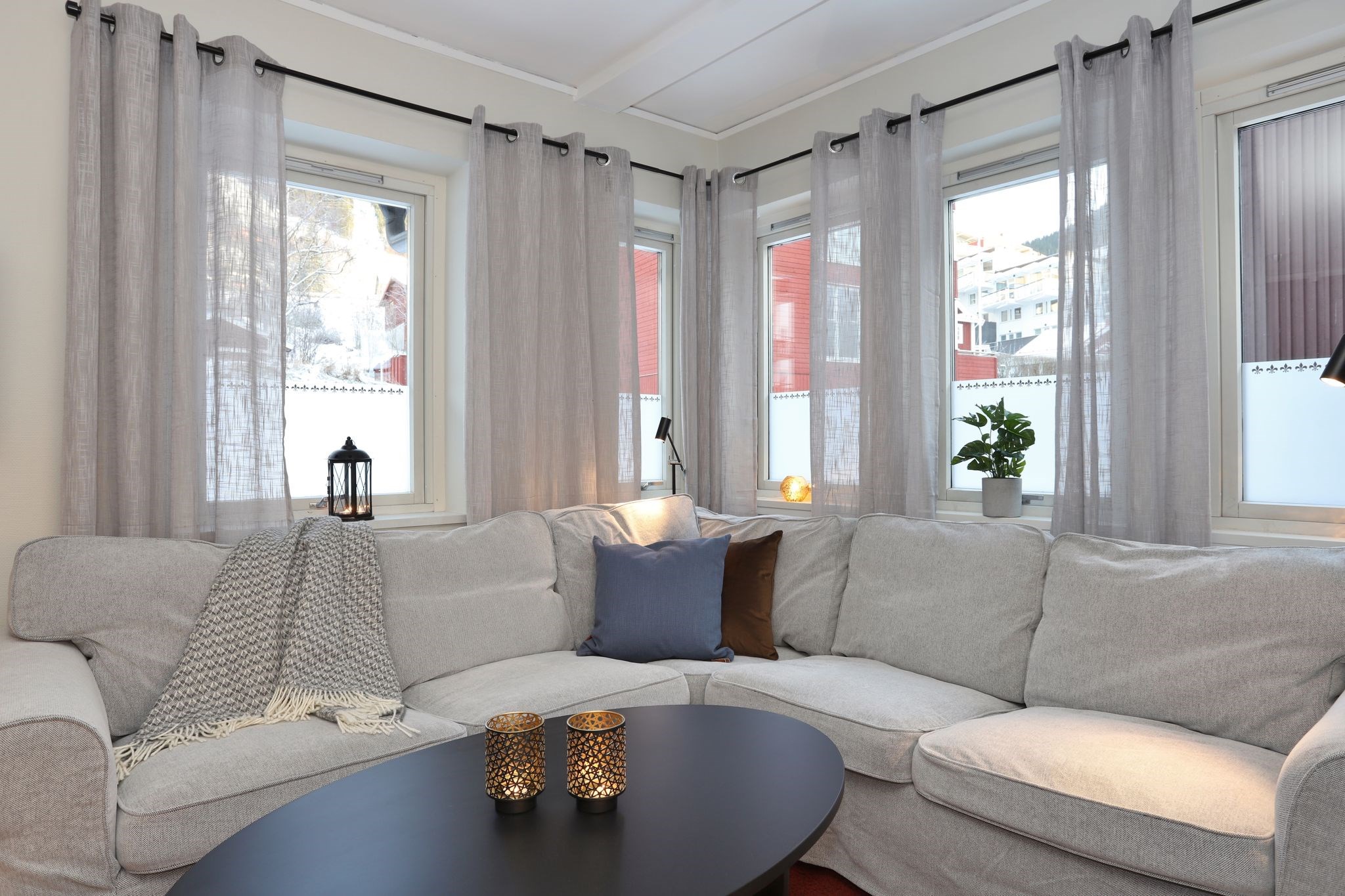 En hyggelig stue med en lysegrå sofa, dekoreret med puder og et tæppe. Et rundt sort sofabord har to lysestager. Store vinduer med tynde gardiner lukker naturligt lys ind.