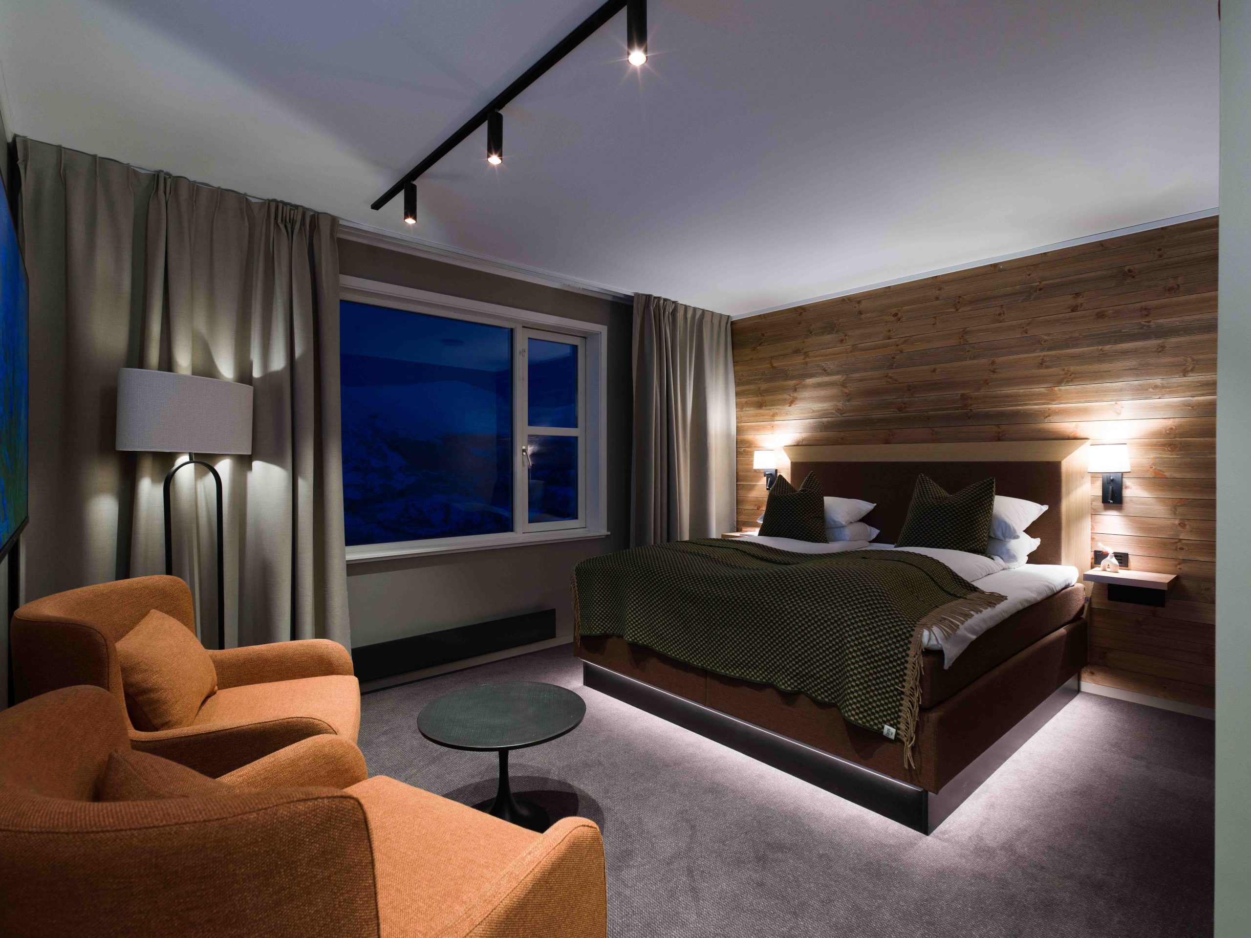 Et moderne hotelværelse med en stor seng, grønt sengetøj, en accentvæg med træpaneler, to orange lænestole, et rundt sort bord, en gulvlampe og et stort vindue med gardiner, der viser et natudsigt udenfor.