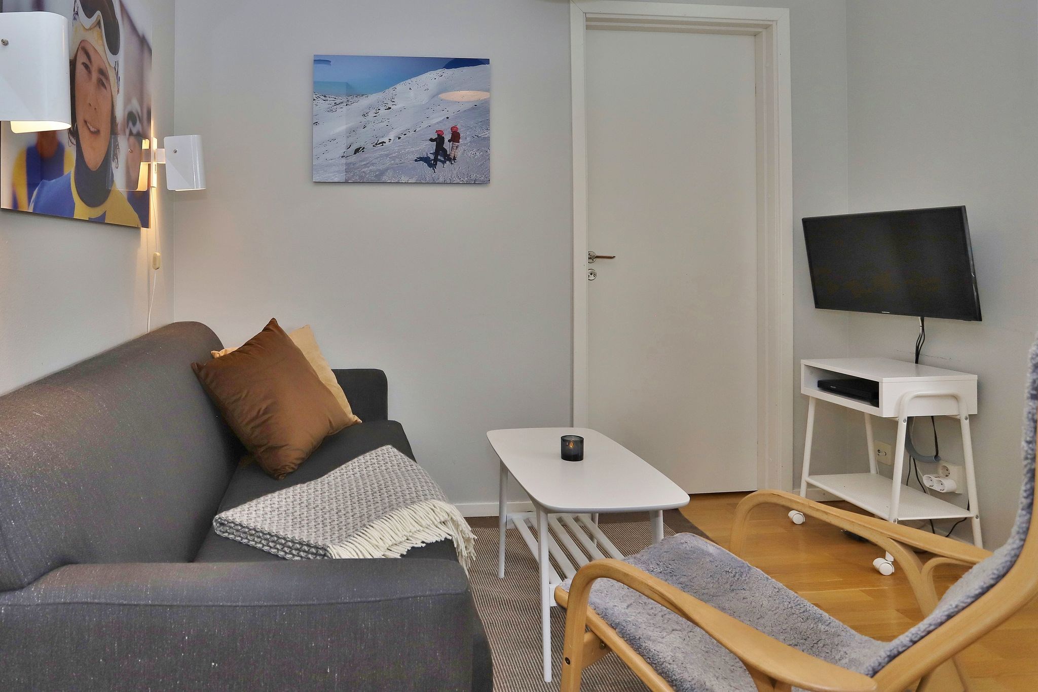 En lille, hyggelig stue med en mørkegrå sofa, brune og grå hynder, et hvidt sofabord, et tv på et hvidt stativ og en grå stol. To landskabs- og portrætfotos hænger på de lysegrå vægge.