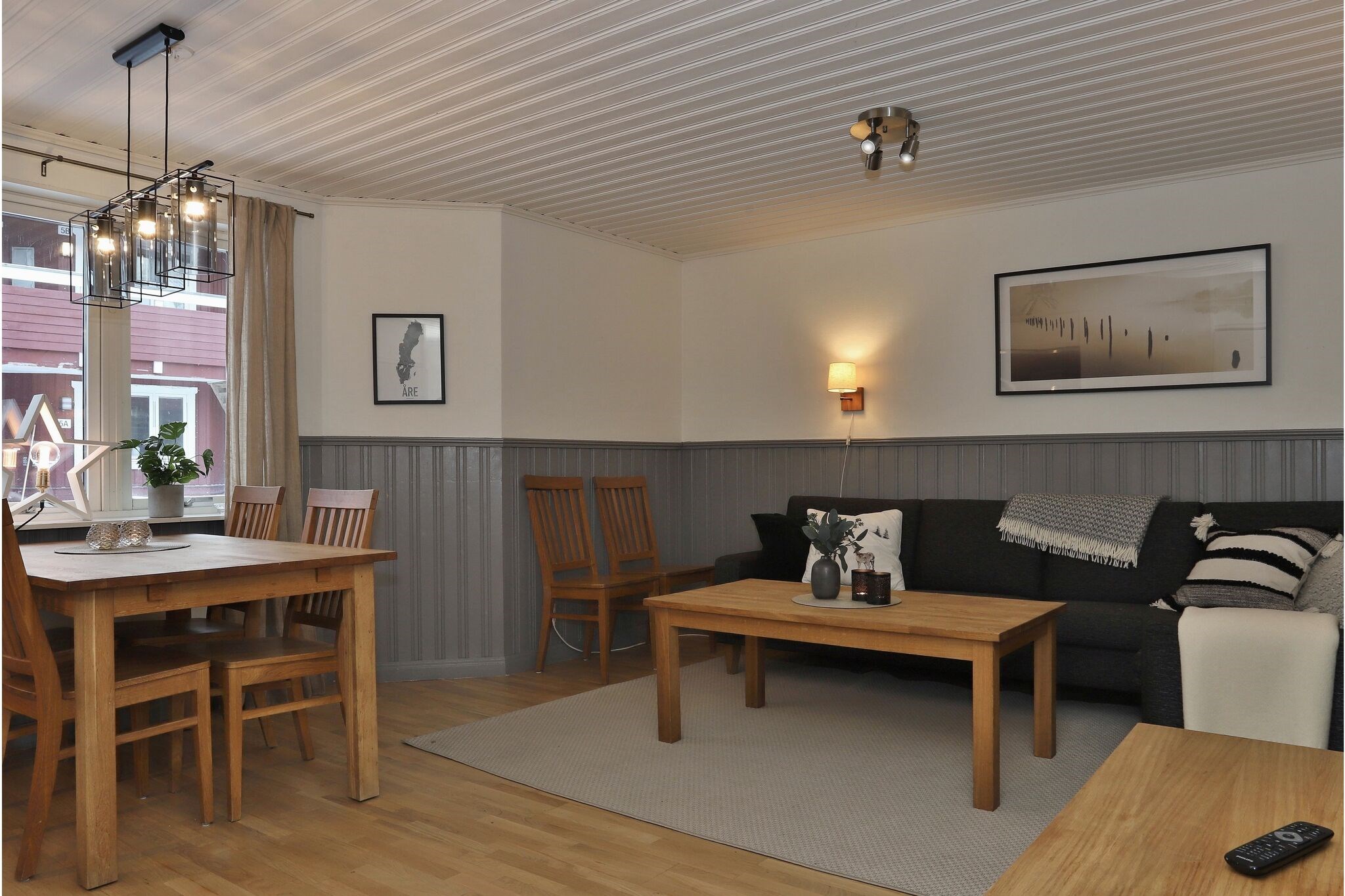 En hyggelig opholds- og spisestue med træmøbler, neutrale vægge, en sort sofa, indrammet kunst på væggene og et stort vindue, der lukker naturligt lys ind. Et spisebord med stole står ved siden af vinduet.