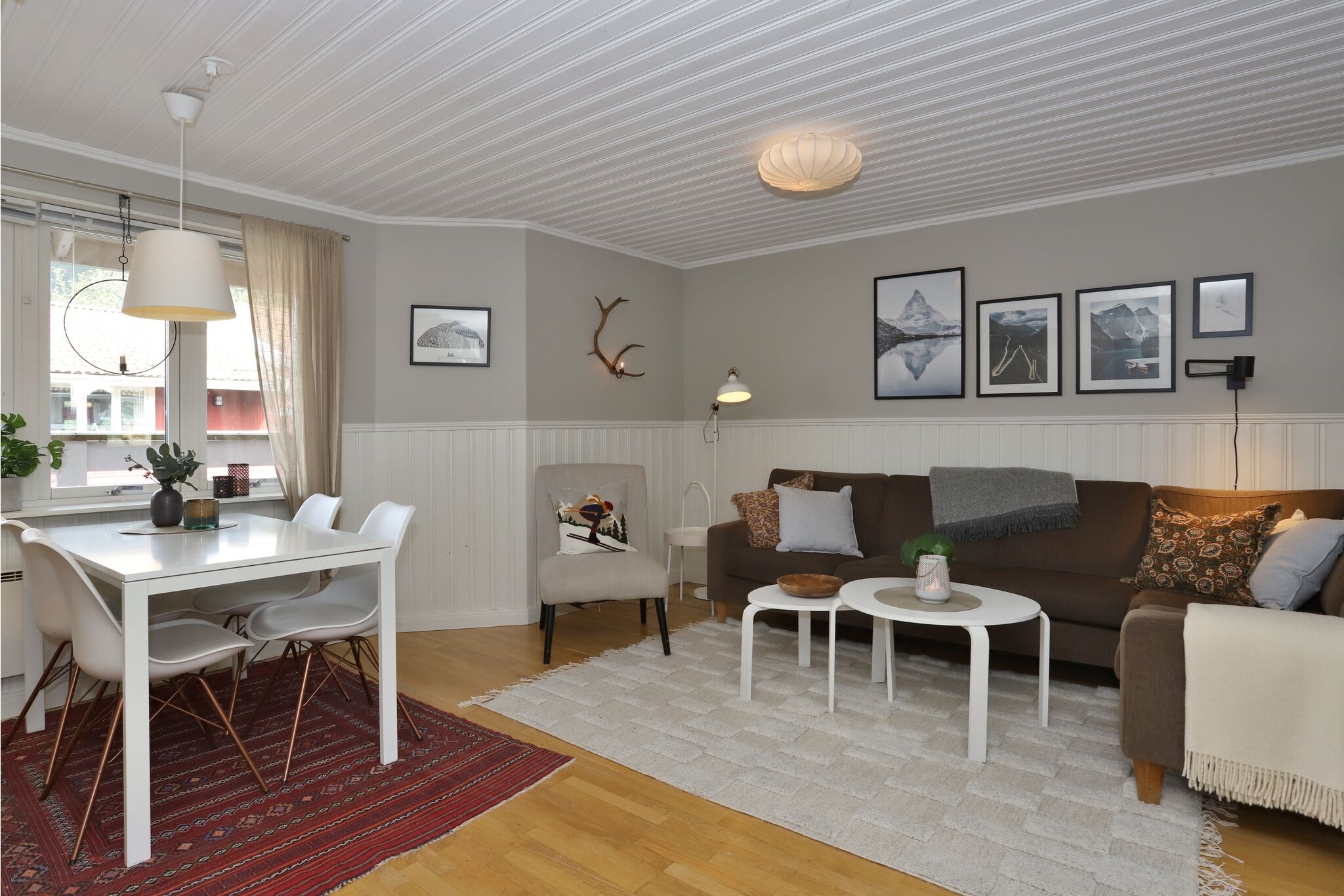 En hyggelig opholds- og spisestue med brun sofa, beige lænestol, hvide borde, spisebordssæt, vægkunst, gevirdekoration og lyst trægulv med et rødt tæppe og et neutralt vævet tæppe. Stort vindue lukker naturligt lys ind.
