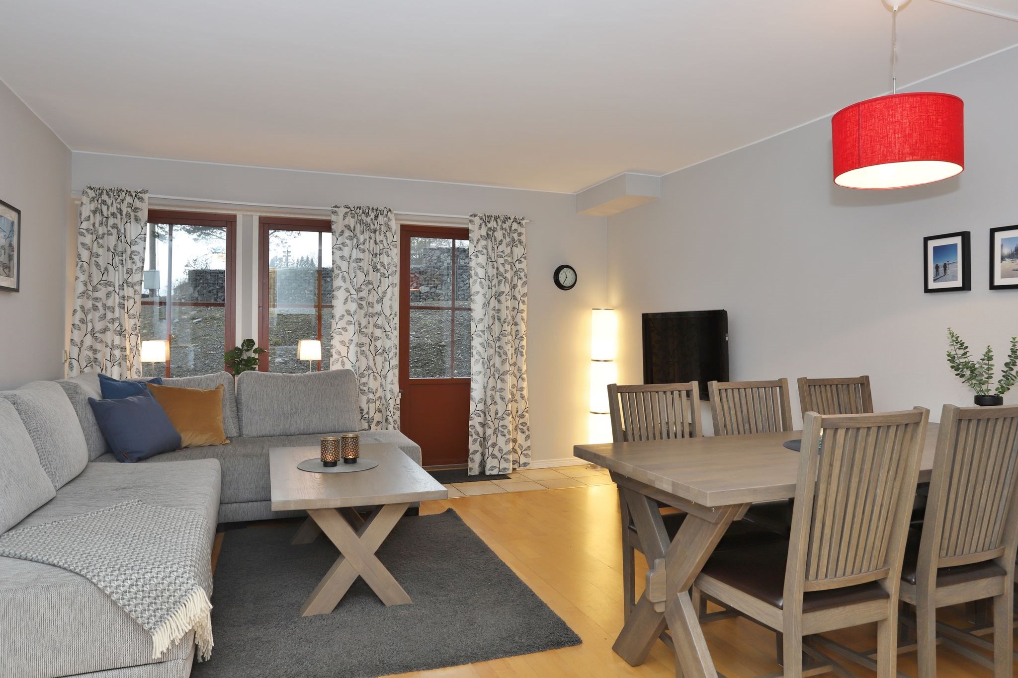 En moderne stue og spisestue med en grå sektionssofa, et spisebord i træ med stole, gulvlampe, vægur, kunstværker og en rød pendellampe. Store vinduer med gardiner lukker naturligt lys ind.