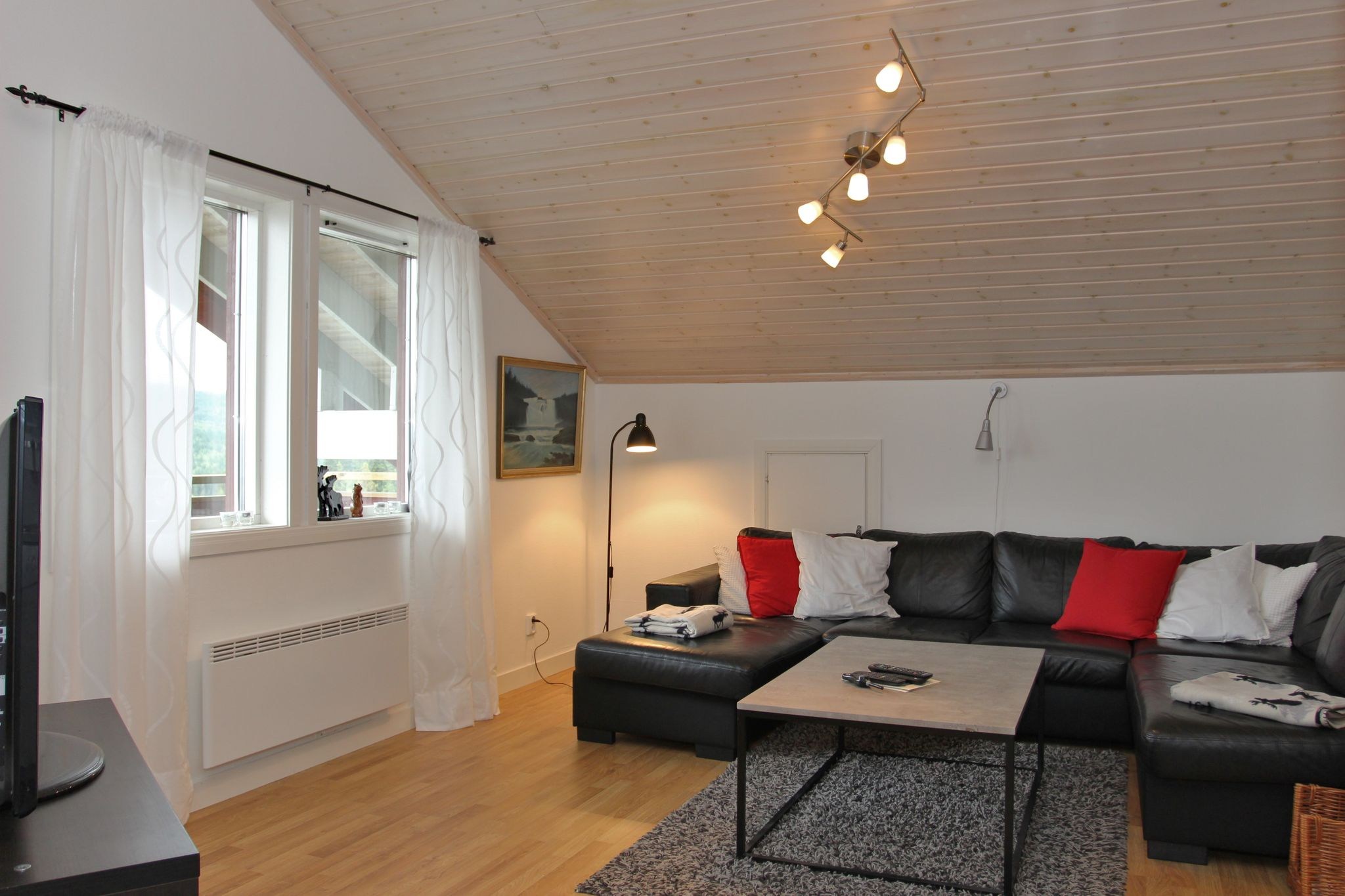 En hyggelig stue med skrånende træloft, sort sofa med røde og hvide hynder, et sofabord, gråt tæppe, gulvlamper og et vindue med tynde gardiner, der lukker naturligt lys ind.