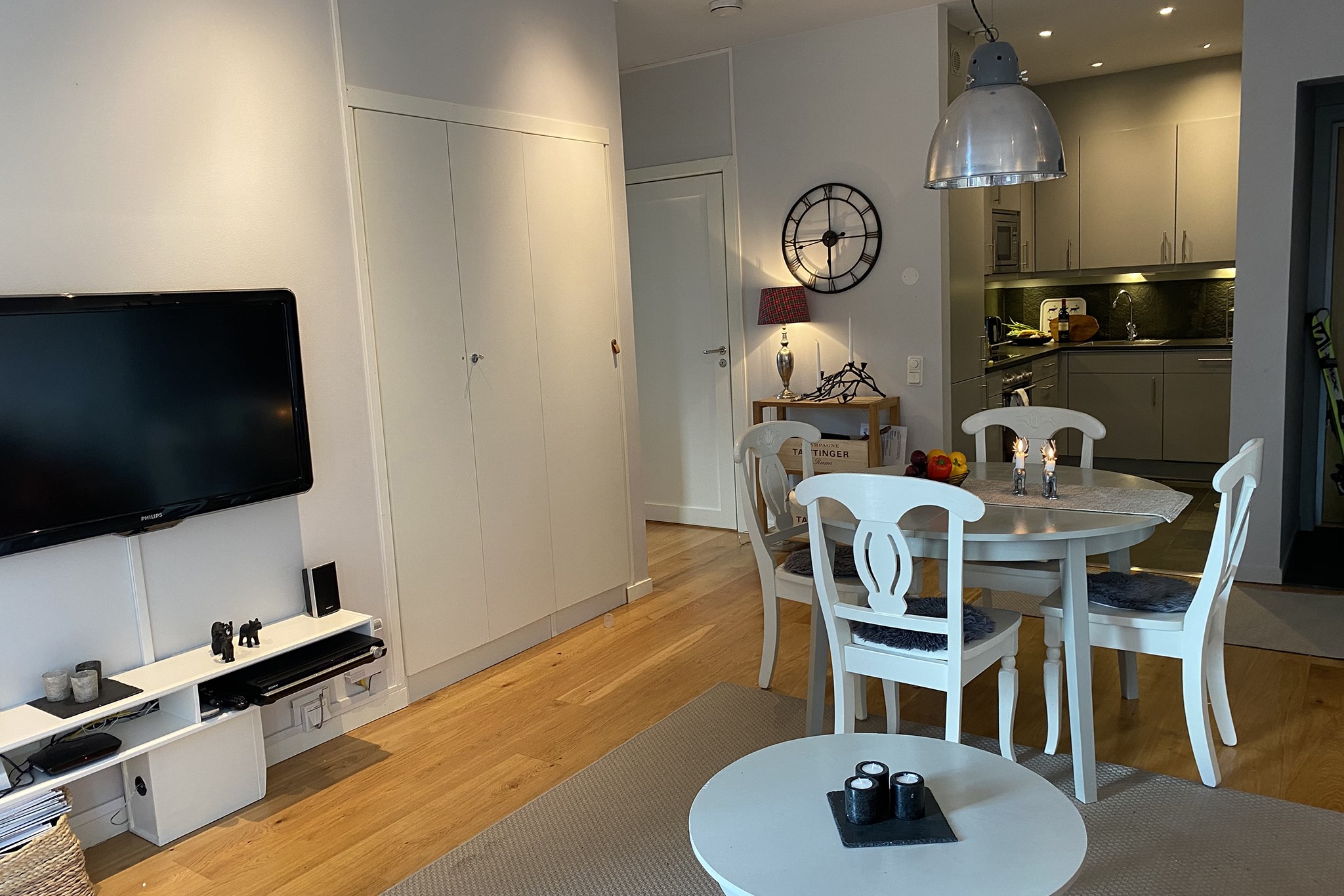 En hyggelig, moderne lejlighed med lyse vægge og trægulve. Der er et rundt spisebord med fire stole, et lille sofabord, et vægmonteret tv og et åbent køkken i baggrunden.