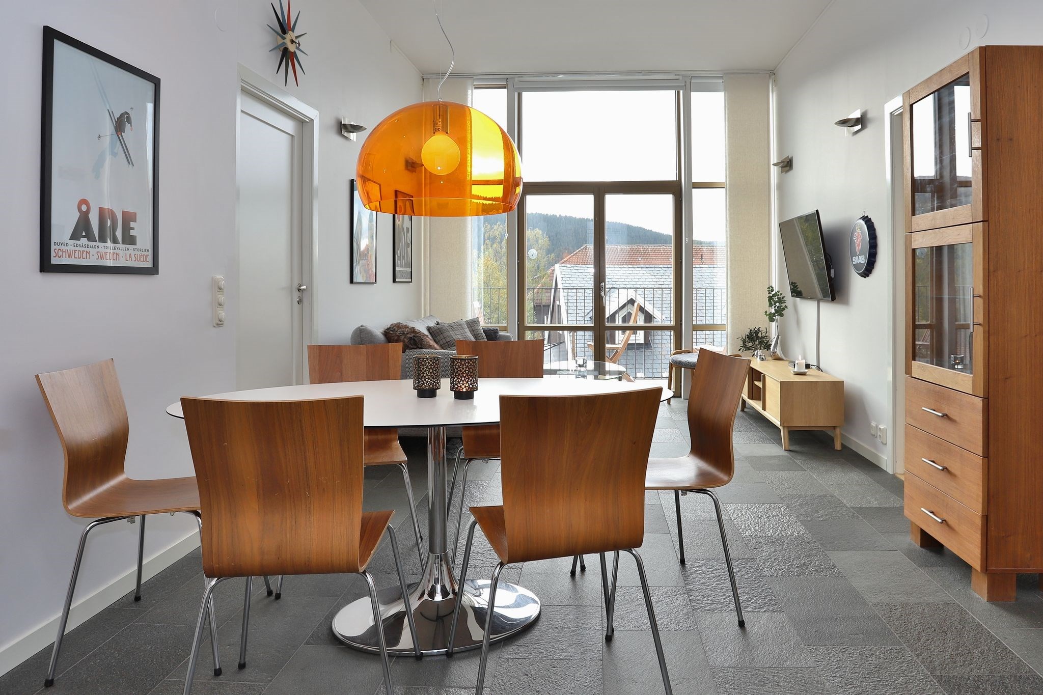 Moderne spise- og opholdsstue med træstole omkring et hvidt bord, en orange hængelampe, et vitrineskab, et vægmonteret tv, store vinduer og kunstværker på hvide vægge. Udsigt over bjergene synlig gennem vinduerne.