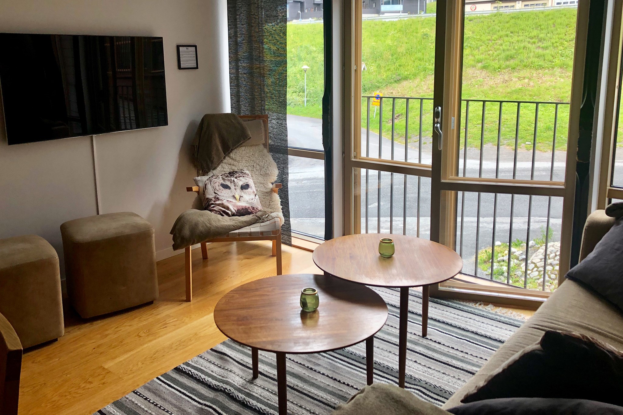 En hyggelig stue med et stribet tæppe, to runde sofaborde i træ, en sofa, en stol med et tæppe og en uglepude, to puffer, et vægmonteret tv og store vinduer med udsigt til græs og en vej udenfor.