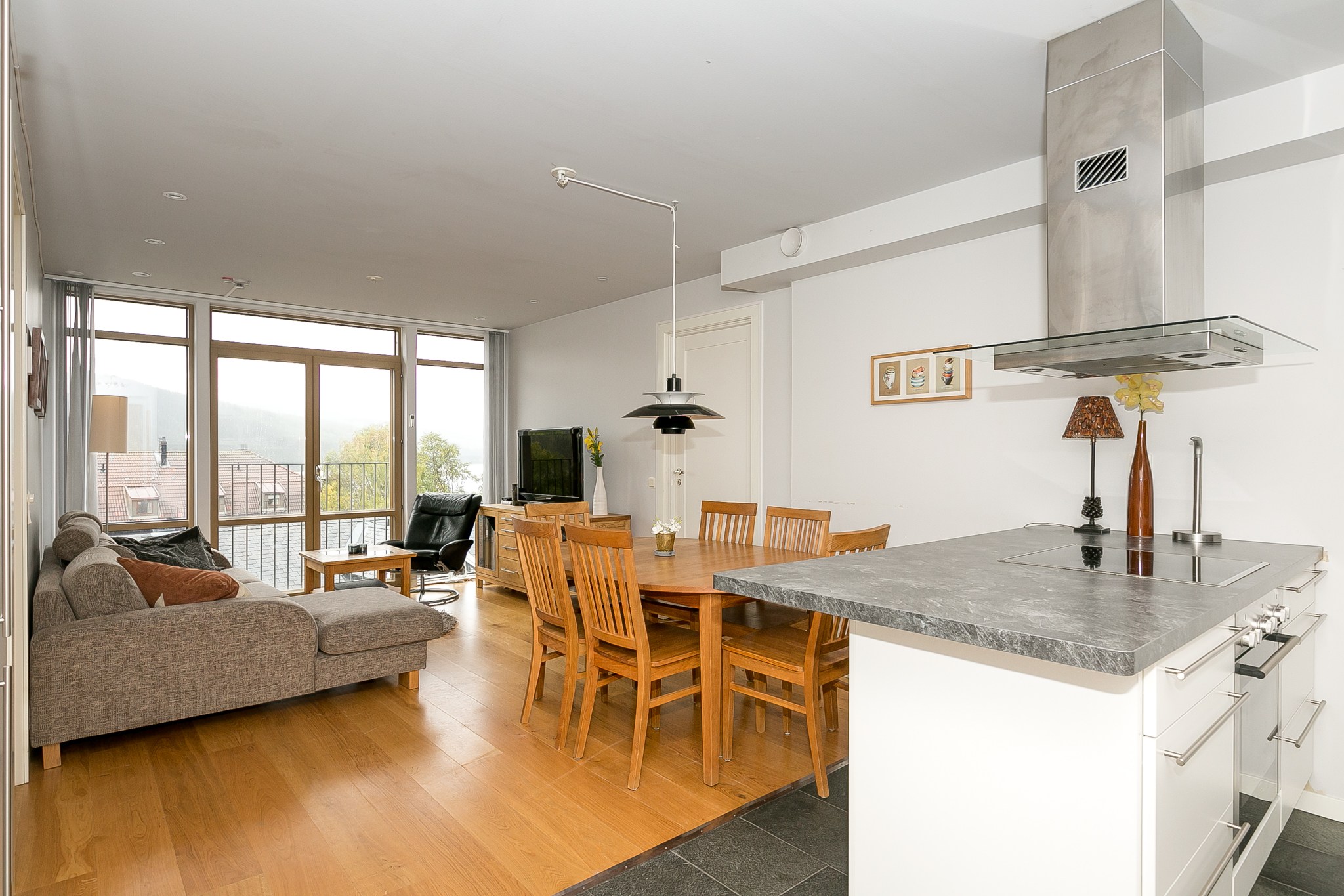 Moderne åben opholdsstue med køkken, spisebord i træ, stole, grå sofa, TV og store vinduer, der lukker naturligt lys ind; lyst trægulv og neutral indretning overalt.