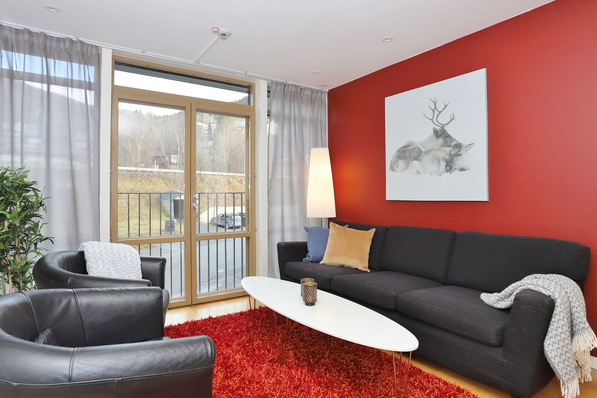 Moderne stue med en rød accentvæg, sort sofa, to sorte lænestole, hvidt ovalt sofabord, rødt shag-tæppe, gulvlampe og stort vindue med tynde gardiner. Et hjortekunstværk hænger over sofaen.