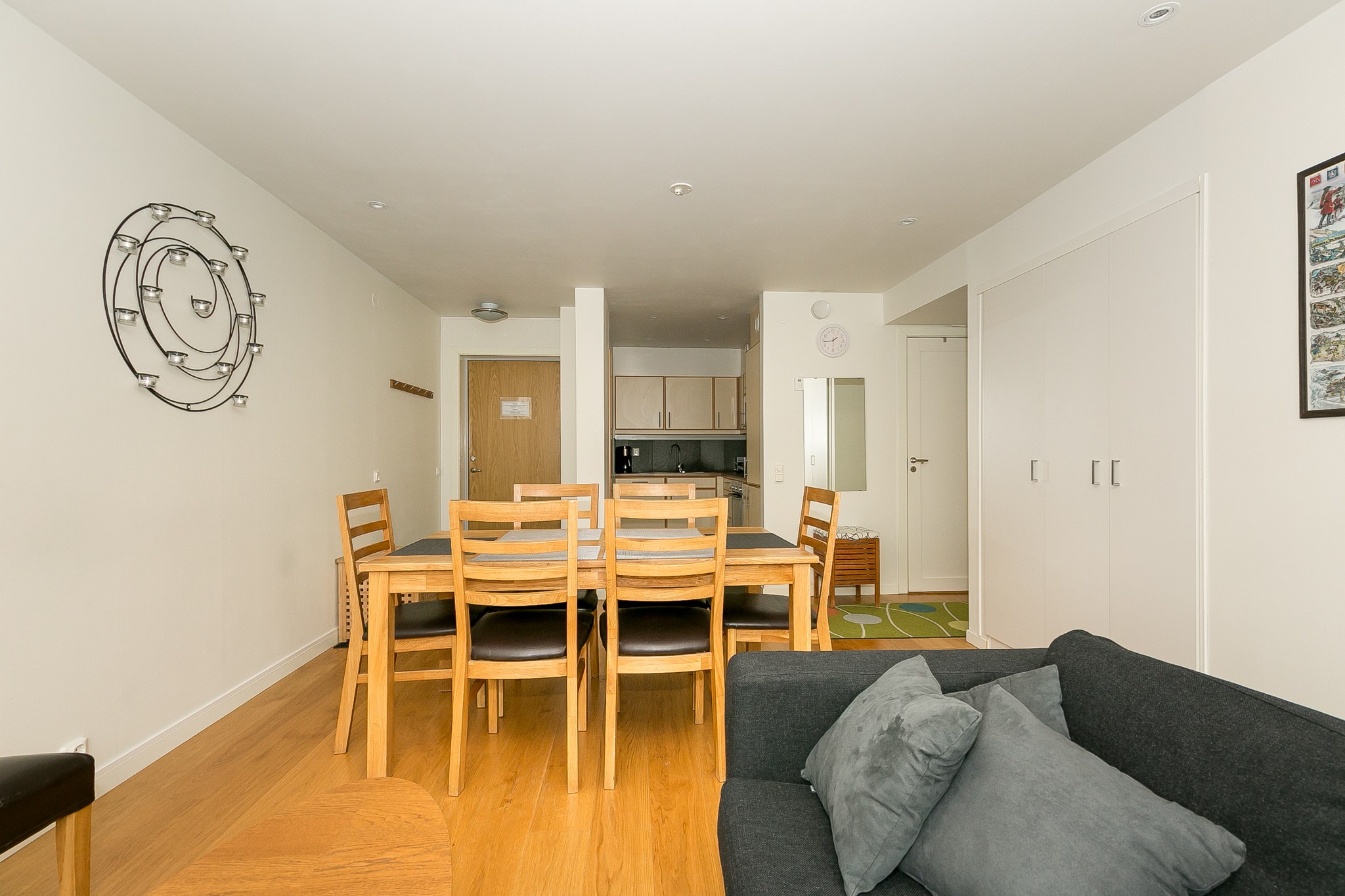 En moderne lejlighedsindretning med et spisebord i træ med seks stole, en sort sofa med hynder, en vægskulptur, et tekøkken i baggrunden og hvide vægge med minimalistisk indretning.