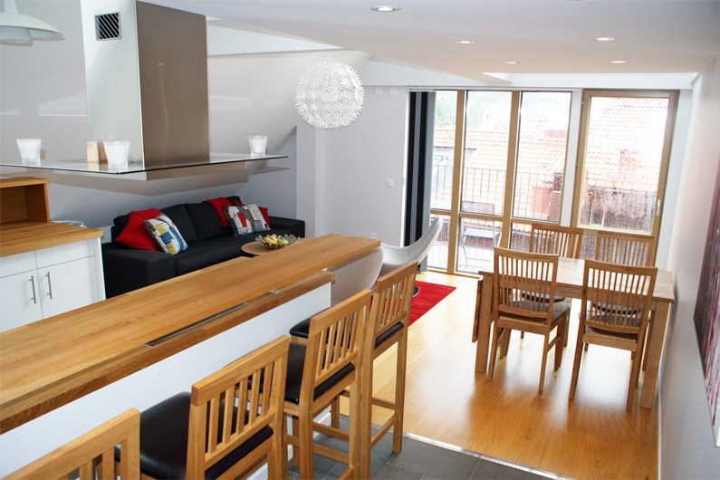 Moderne åbent køkken og opholdsstue med trægulve, morgenmadsbar med taburetter, spisebord, sofa med farverige hynder og store vinduer, der lukker naturligt lys ind.