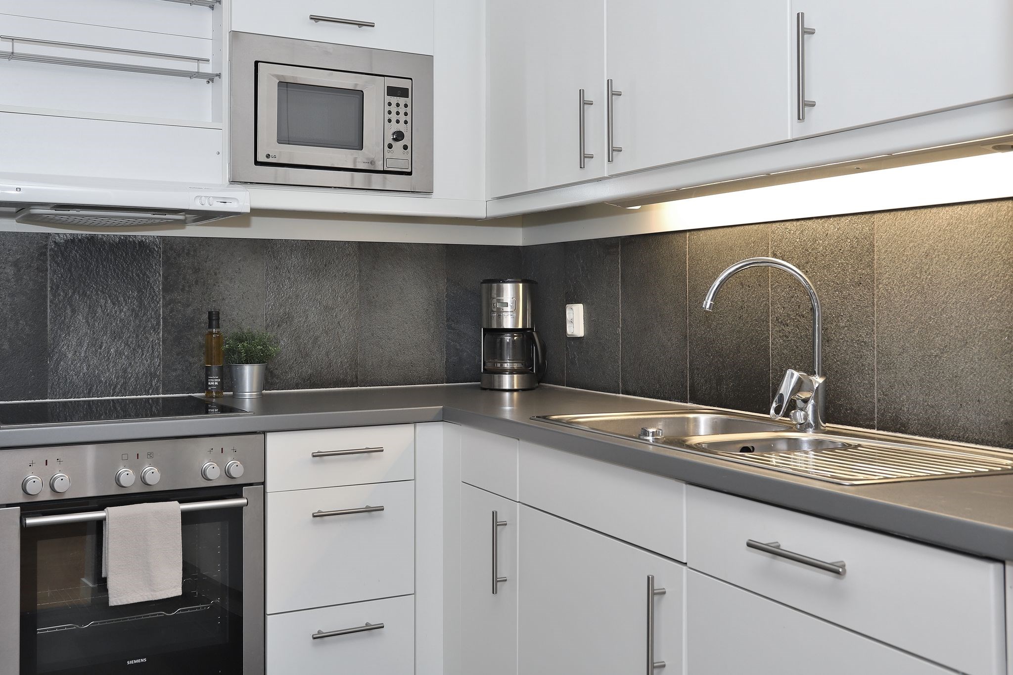 Moderne køkken med hvide skabe, indbygget ovn, mikrobølgeovn, kaffemaskine, vask i rustfrit stål, belysning under skabet og sort flisebelagt bagplade. En lille plante og en flaske olivenolie står på køkkenbordet.