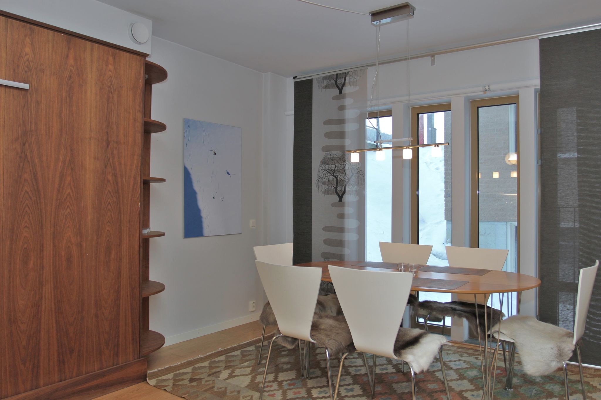 Moderne spisestue med et træbord, seks hvide stole med pelsdække, mønstret tæppe, stort træskab, abstrakt vægkunst og gulv-til-loft-vinduer med tynde grå gardiner.