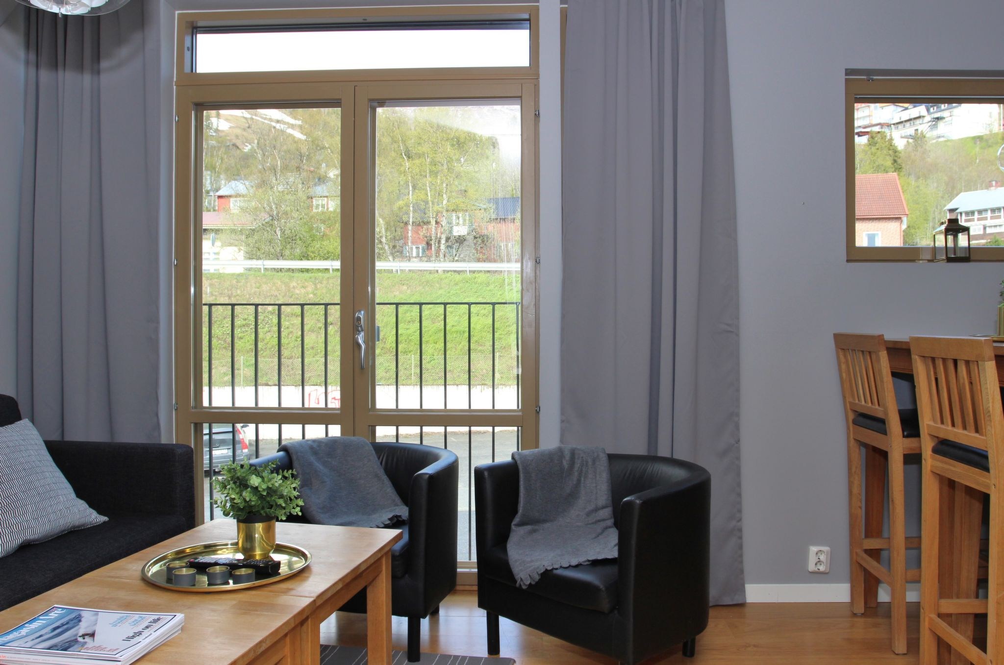 Moderne stue med store glasdøre, der fører ud til en balkon, to sorte lænestole med grå tæpper, et sofabord i træ med bakke og plante og høje stole ved en morgenmadsbar nær et spejl på en grå væg.