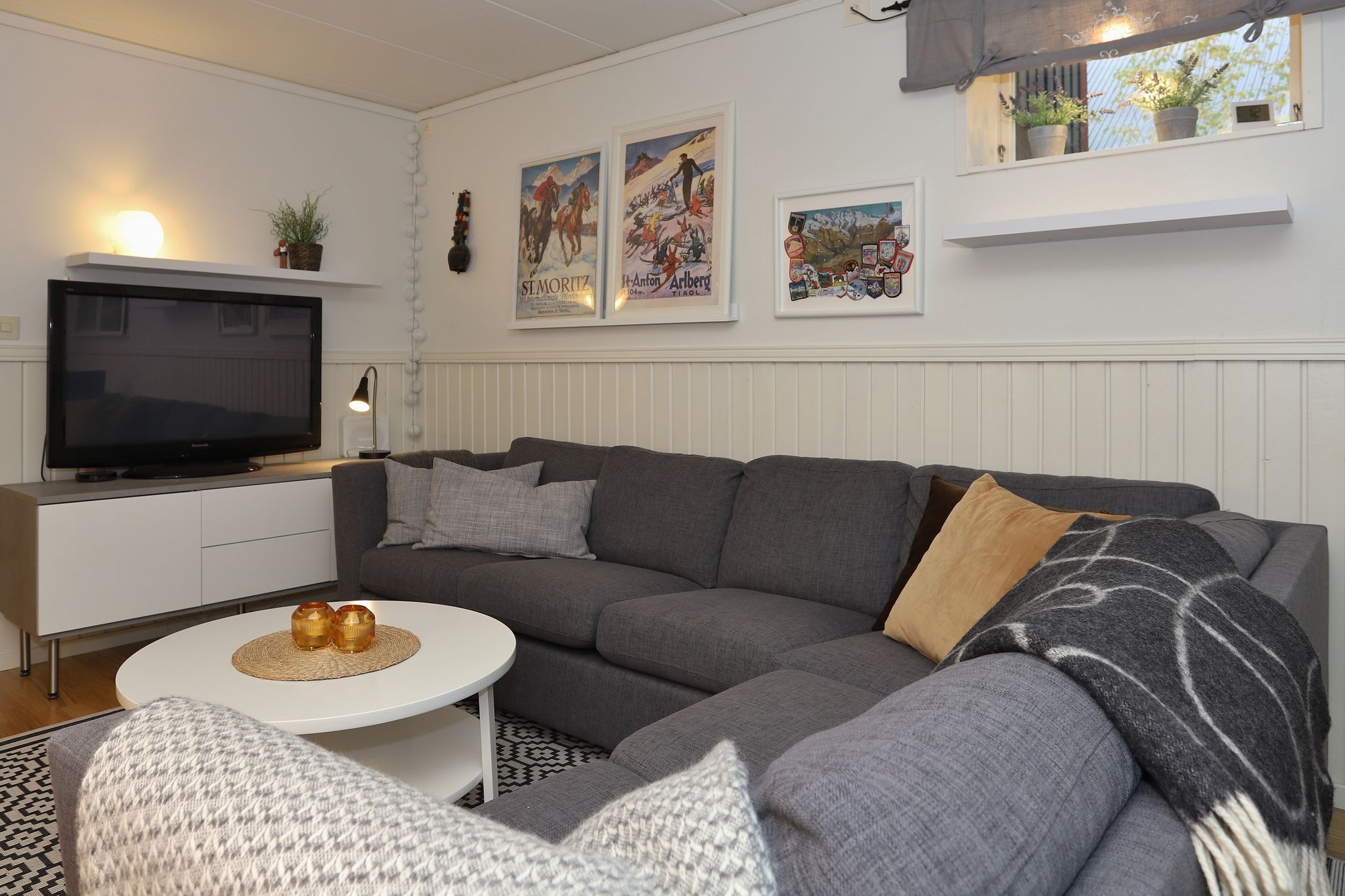 En hyggelig stue med en grå sofa, forskellige puder, et rundt hvidt sofabord med stearinlys, et tv på et hvidt stativ, vægkunst og et lille vindue med en hylde med planter.