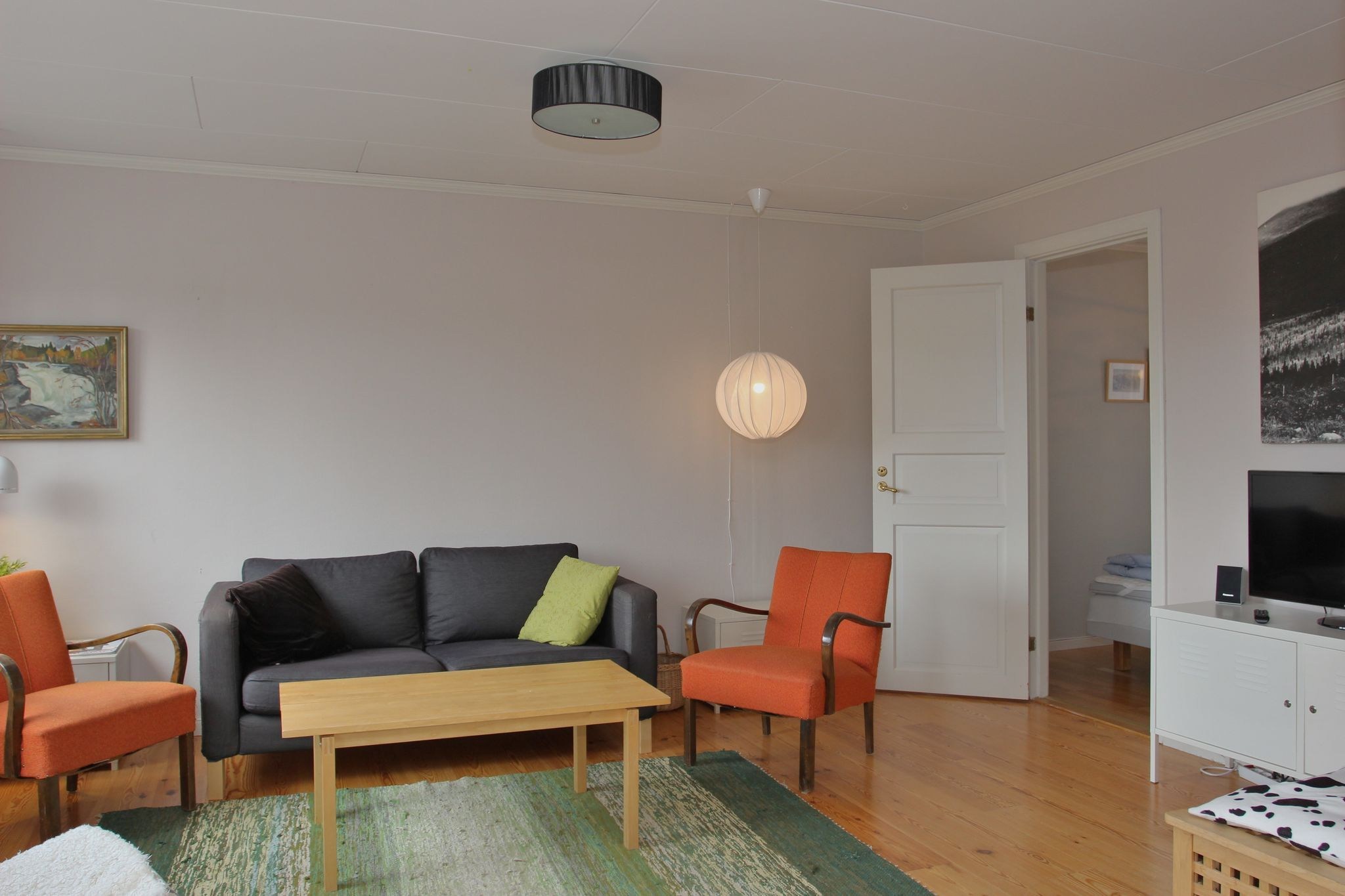 En hyggelig stue med en sort sofa, to orange lænestole, et sofabord i træ og et grønt tæppe. En rund hængelampe, et tv og en åben dør til et soveværelse er også synlige. Gulvet er af træ, og der hænger kunstværker på væggen.
