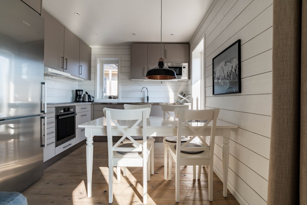 Et moderne køkken med lyse trægulve, hvide vægge med kant og lysegrå skabe. Et hvidt spisebord med to stole står under en sort pendellampe. Sollys strømmer ind gennem vinduet og skaber en lys og hyggelig atmosfære.