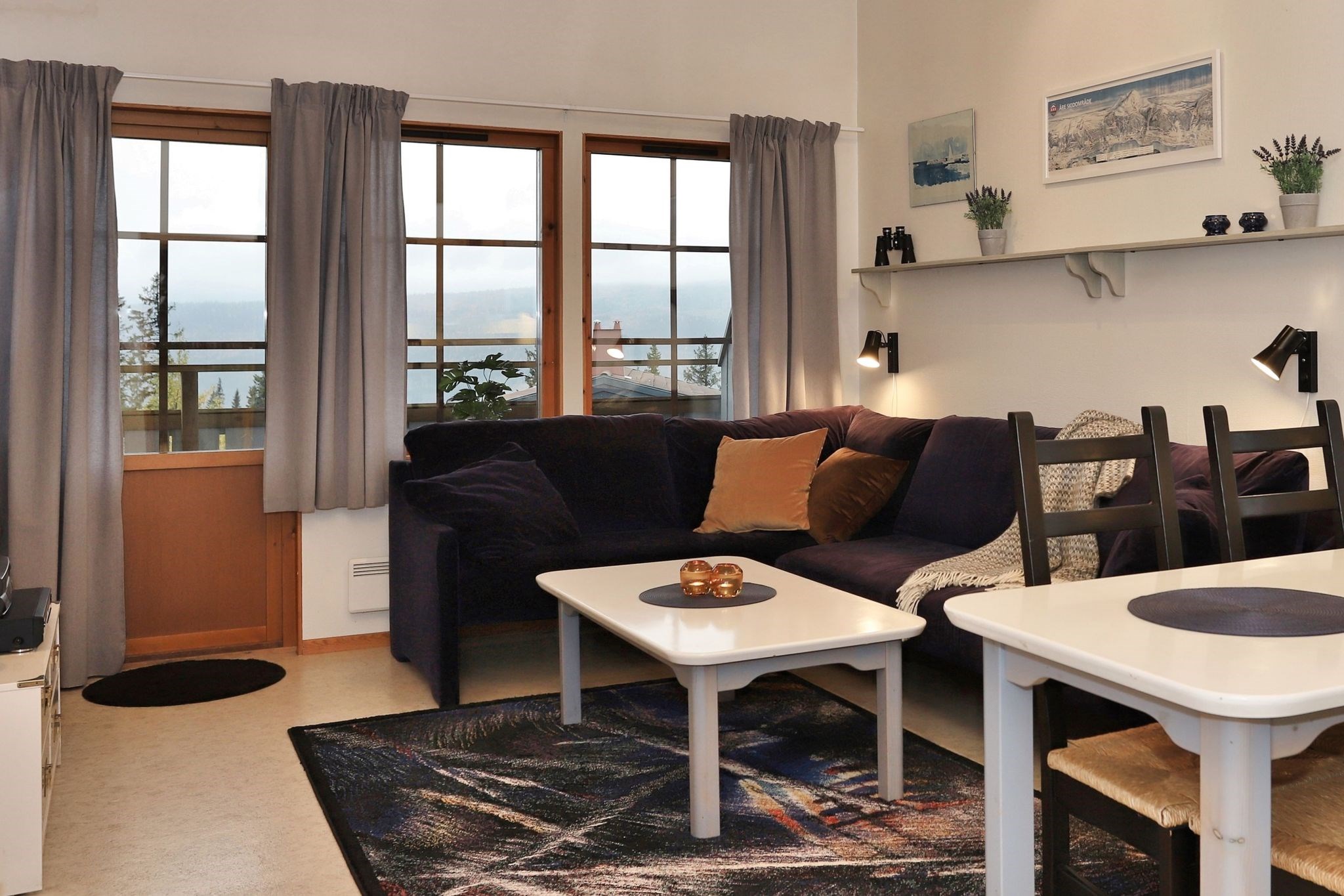 Hyggelig stue med en mørk L-formet sofa, farverige hynder, et sofabord og spiseplads. Store vinduer med grå gardiner lukker naturligt lys ind. Væghylder udstiller små planter og indrammet kunst.