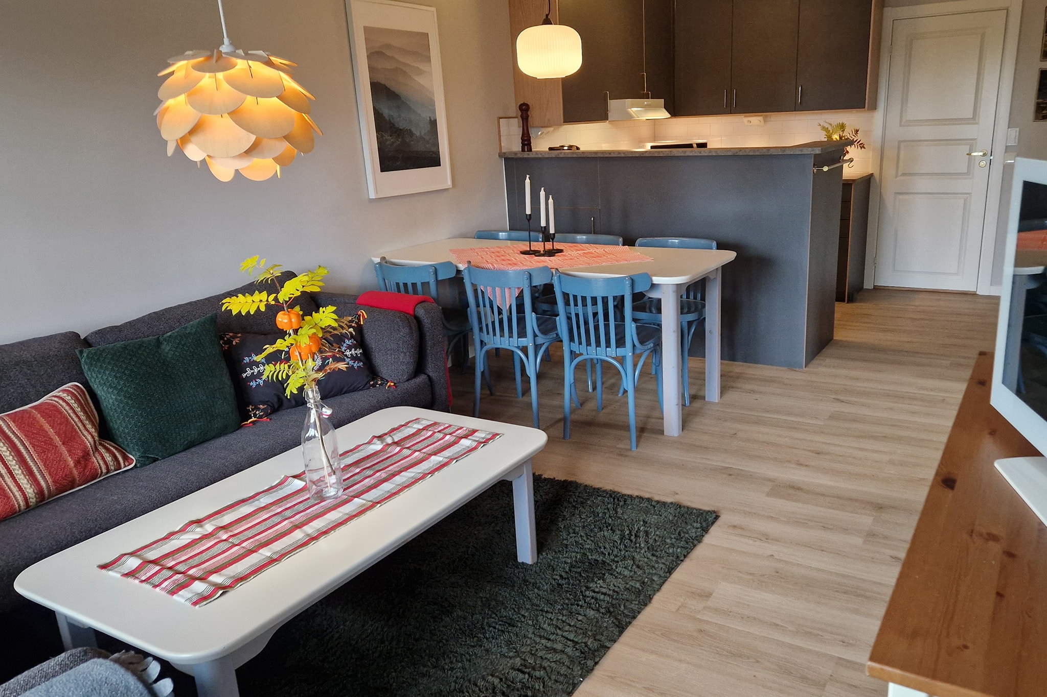 En hyggelig stue og spisestue med en grå sofa, farverige hynder, et hvidt sofabord med en vase med gule blomster, et spisebord med blå stole og et moderne køkken i baggrunden.