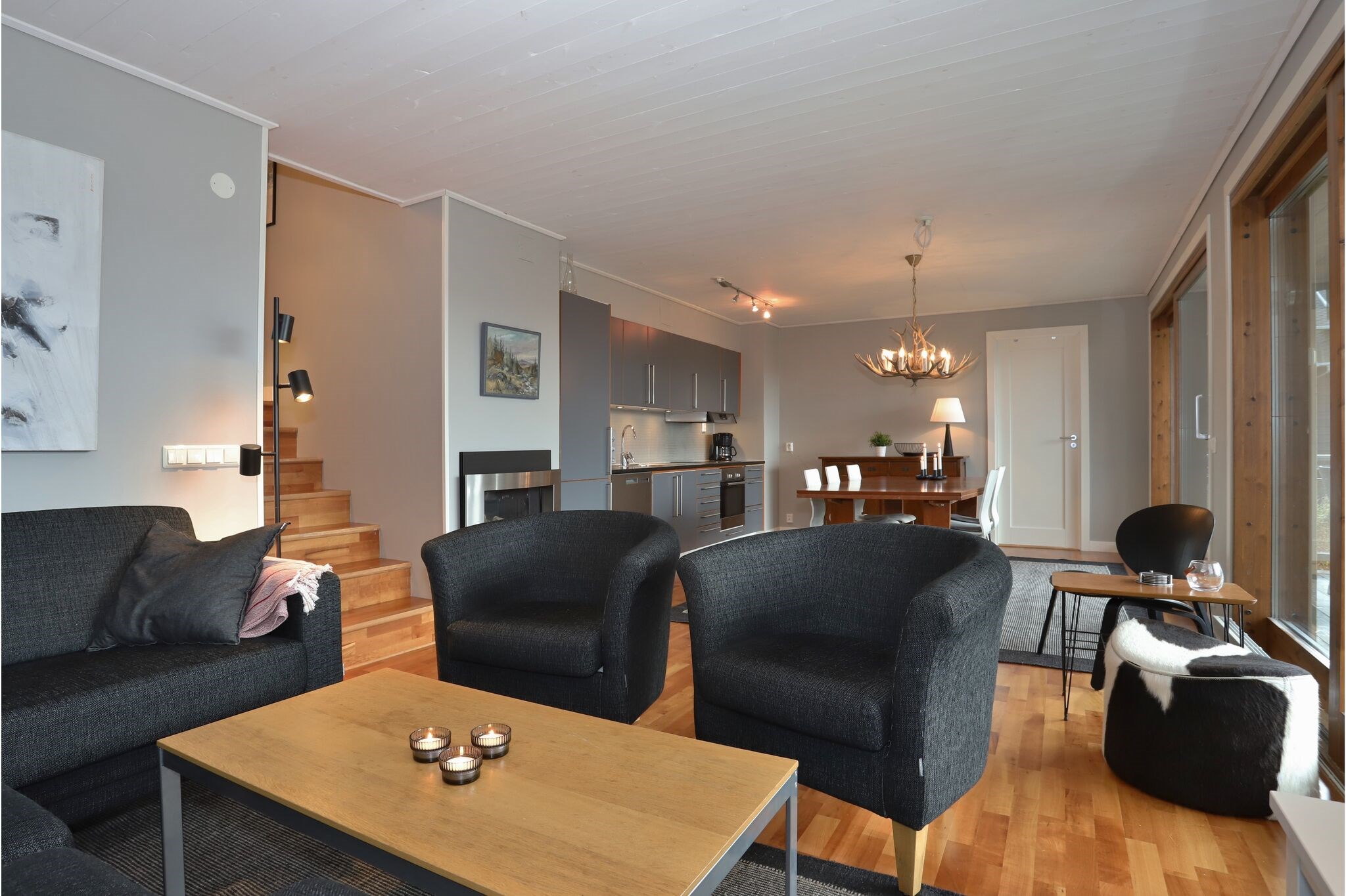 Moderne stue med mørke sofaer og stole, et sofabord i træ med stearinlys, åbent køkken, spiseplads, trægulve og store vinduer, der lukker naturligt lys ind. Trappe fører til en øvre etage.