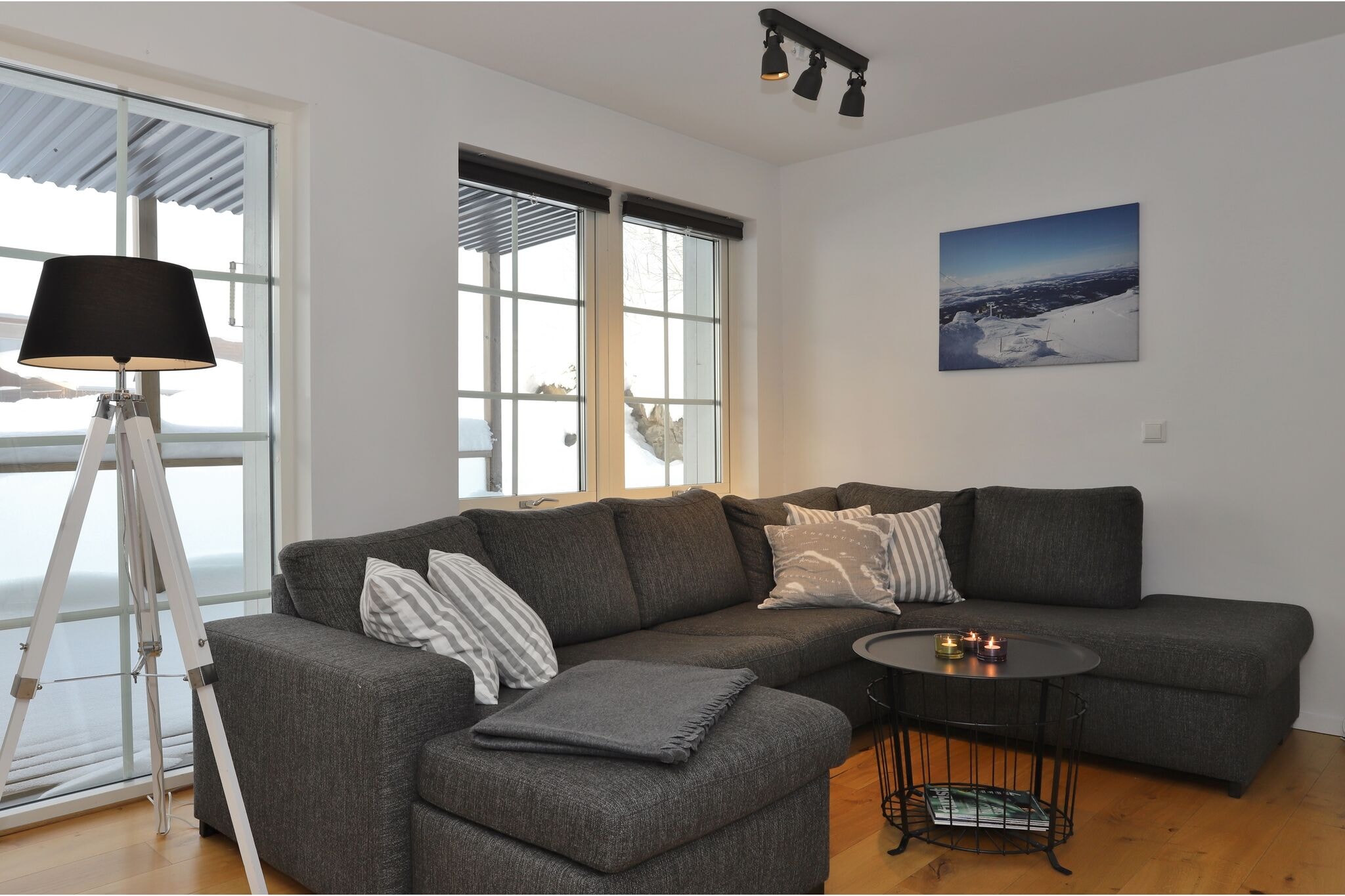 En moderne stue med en mørkegrå sofa, stribede puder, et tæppe, et rundt sort sofabord med stearinlys, en trebenet gulvlampe, store vinduer og et bjergfoto på væggen.