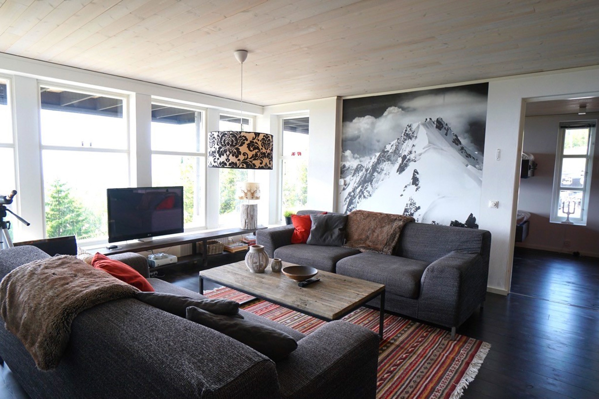 En moderne stue med store vinduer, to grå sofaer, et stribet tæppe, et tv og et sofabord. Et stort sort-hvidt bjergmaleri dekorerer væggen bag den ene sofa.