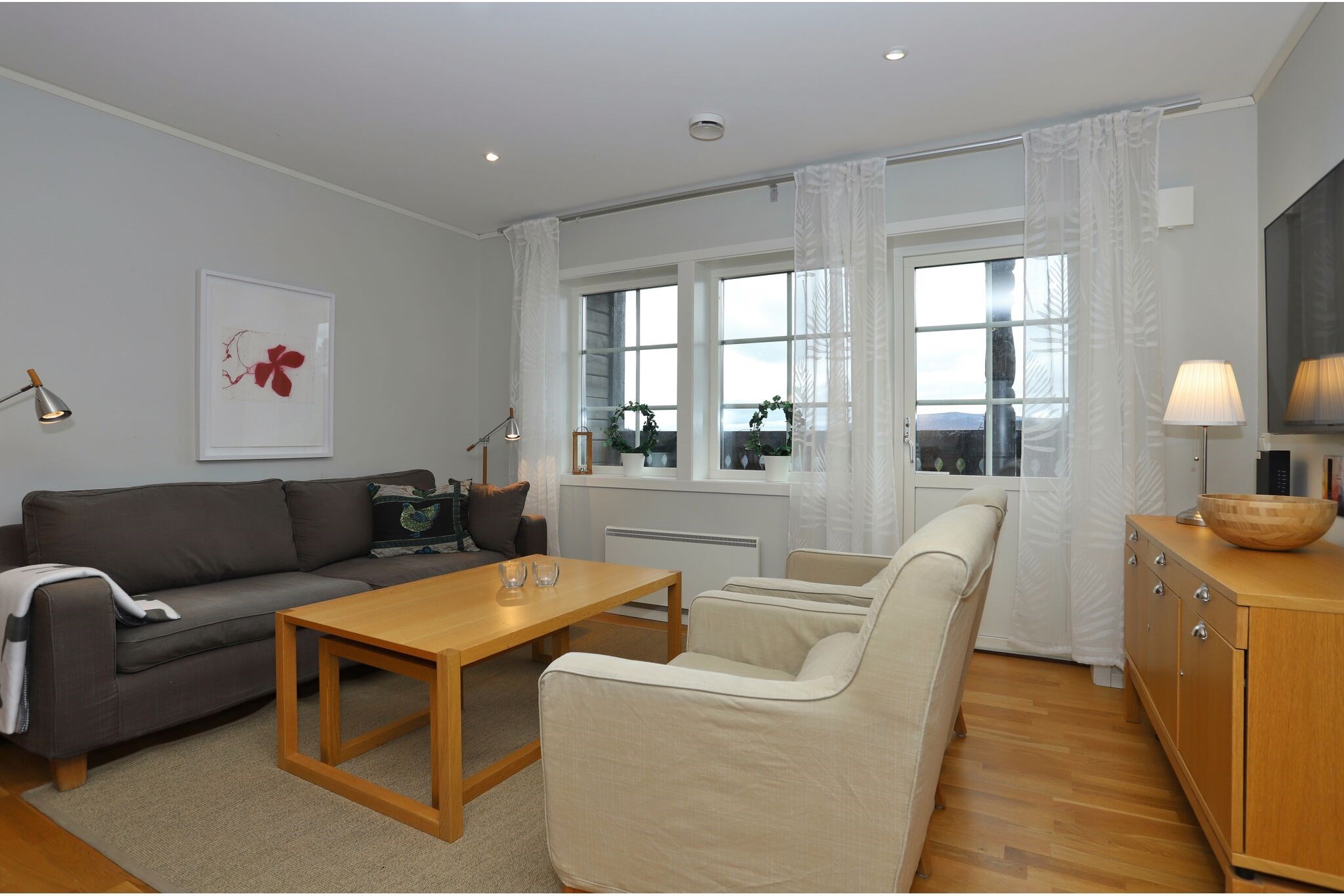 En moderne stue med en grå sofa, to beige lænestole, et sofabord i træ og en skænk med en lampe og skål. Store vinduer med tynde gardiner lukker naturligt lys ind. Et kunstværk med røde blomster hænger på væggen.