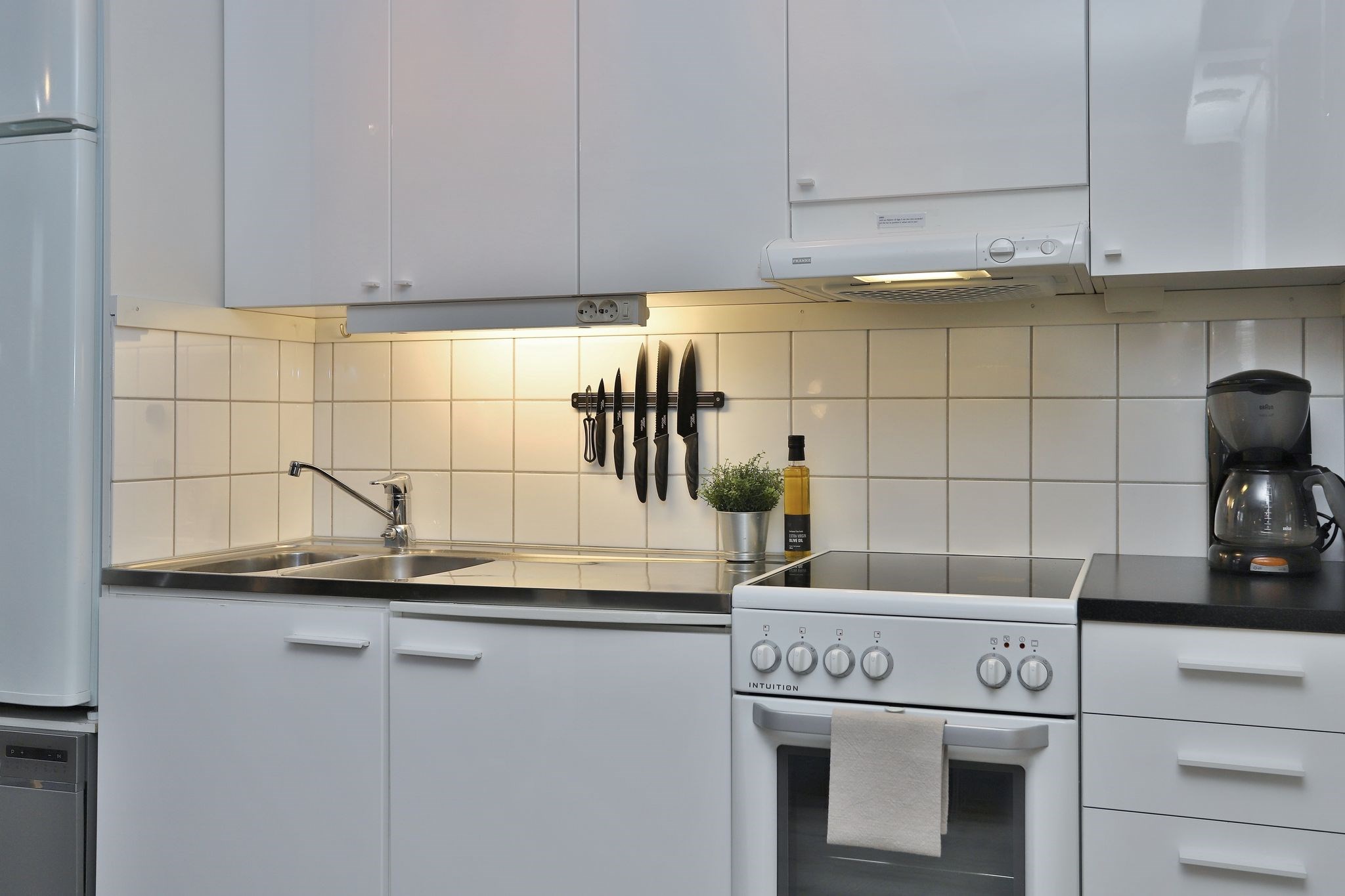 Moderne køkken med hvide skabe, en vask, komfur med håndklæde, kaffemaskine, potteplante, flaske og en magnetisk knivliste med seks knive på en flisebelagt bagplade under skabsbelysning.