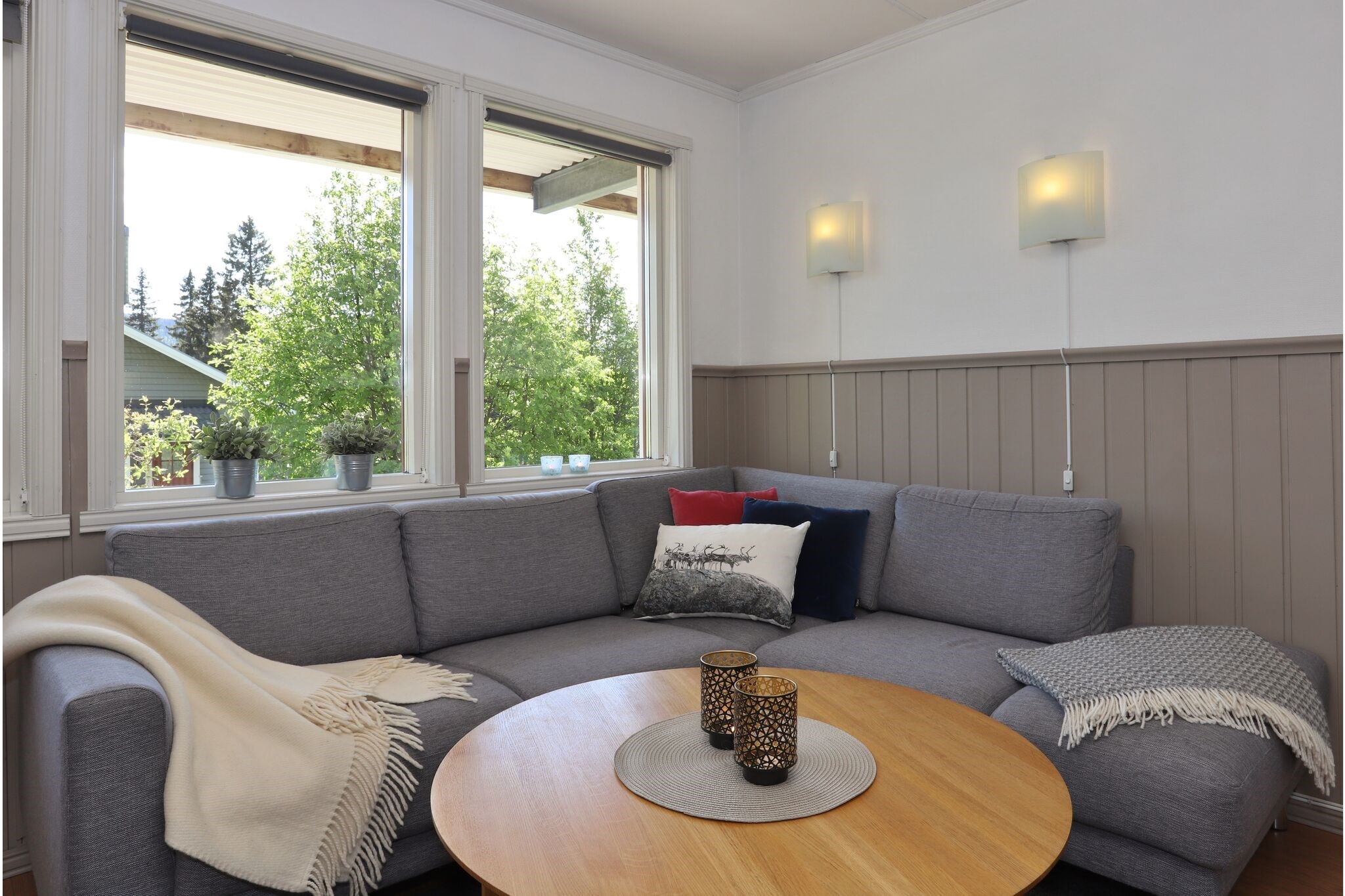En hyggelig stuehjørne med en grå sofa, tæpper og dekorative puder. Et rundt sofabord i træ har plads til to lysestager. Store vinduer bringer naturligt lys ind og viser træerne udenfor.