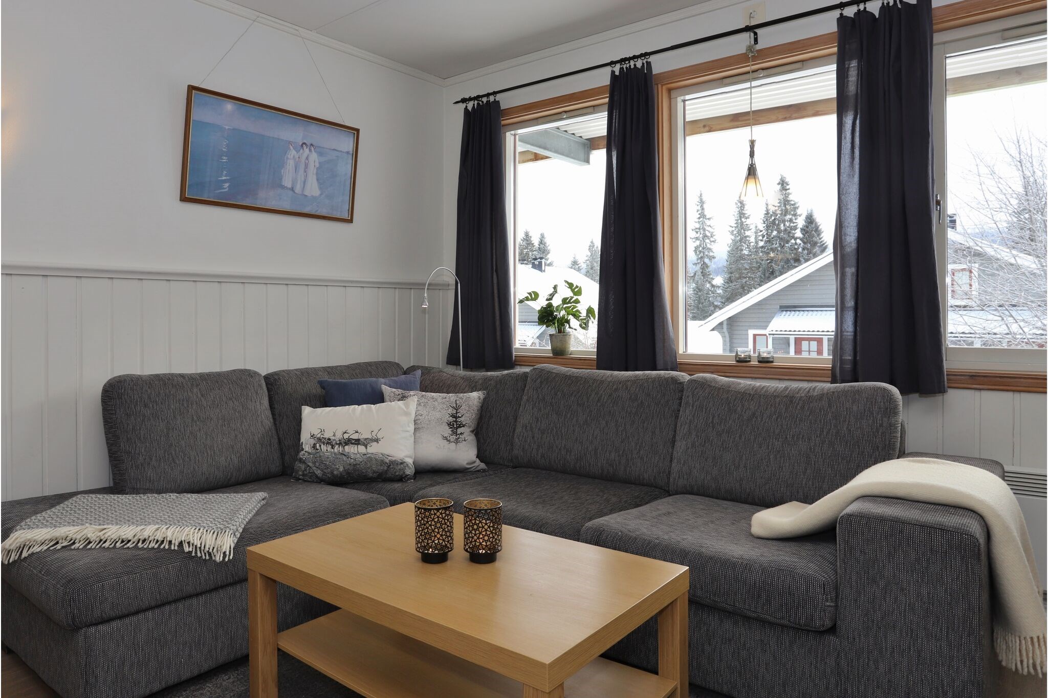 En hyggelig stue med en grå sofa, puder, et tæppe, et sofabord i træ med stearinlys, gardiner for store vinduer og et indrammet billede på en hvidpaneleret væg. Udenfor kan man se træer og et hus.