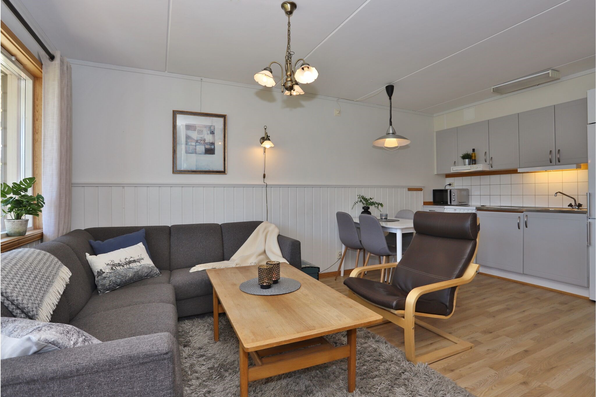 En hyggelig stue og køkkenalrum med grå sofaer, et sofabord i træ, en brun lænestol, et spisebord med fire stole, lysegrå skabe og trægulve, dekoreret med kunst og planter.