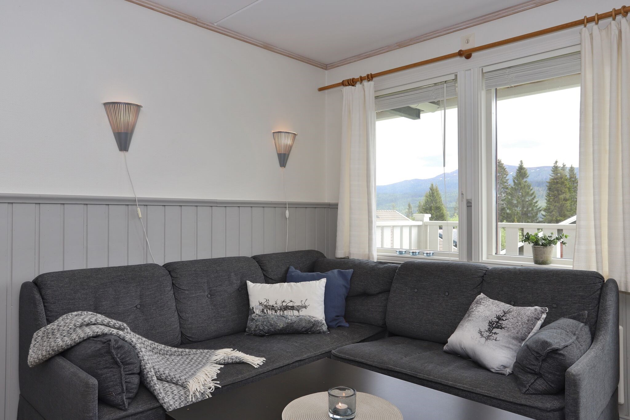 En hyggelig stue med en mørkegrå sofa, dekorative puder, et mønstret tæppe, væglampetter, hvide gardiner og et sofabord med et stearinlys. Store vinduer har udsigt over træer og bjerge.