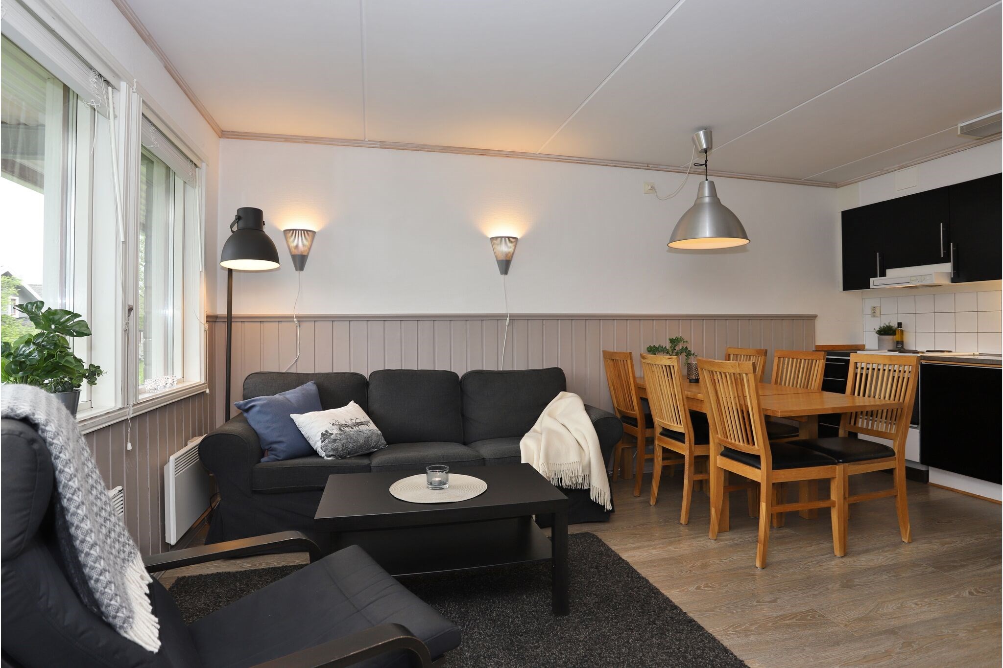 En hyggelig opholds- og spisestue med en sort sofa, lænestol, spisebord i træ med seks stole, moderne belysning og et lille køkken med sorte skabe og hvide fliser. Stort vindue bringer naturligt lys ind.