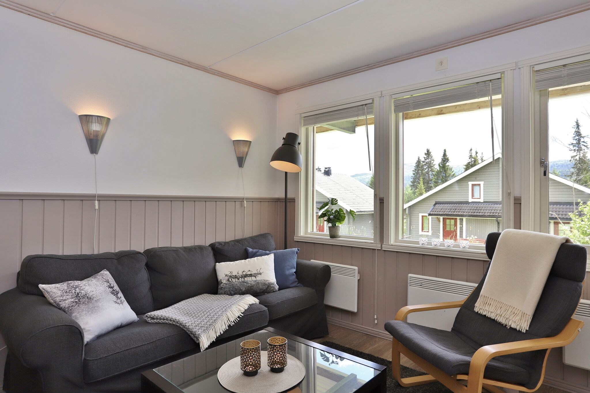 En hyggelig stue med en mørk sofa, lænestol og et tæppe, ved siden af store vinduer med udsigt til huse og træer udenfor. Væglamper og en sort gulvlampe giver blød belysning indendørs.