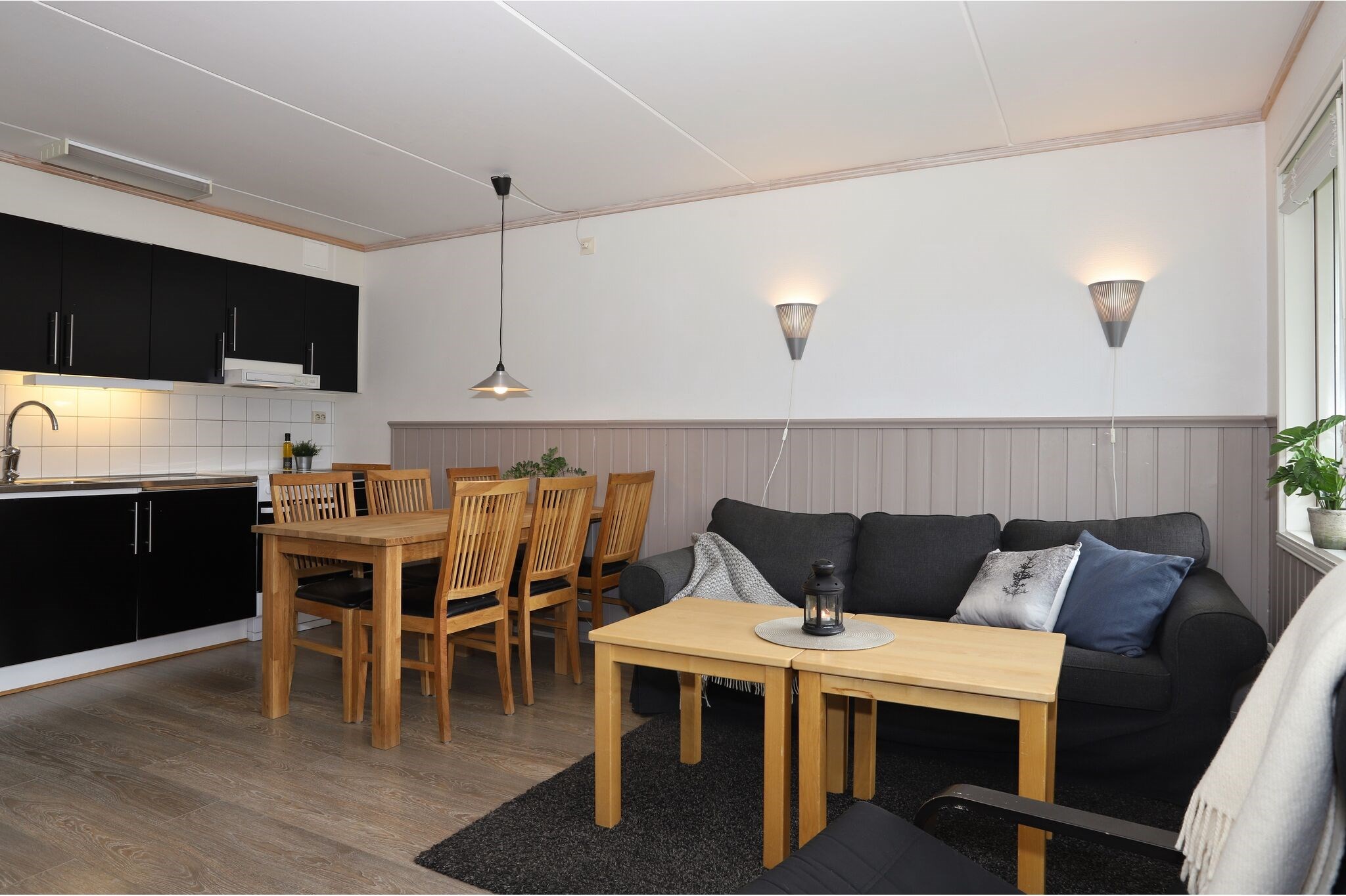 En hyggelig opholds- og spisestue har en sort sofa med hynder, et sofabord i træ, et spisebord med seks stole, sorte køkkenskabe og vægmonterede lamper, alt sammen i en moderne, minimalistisk stil.