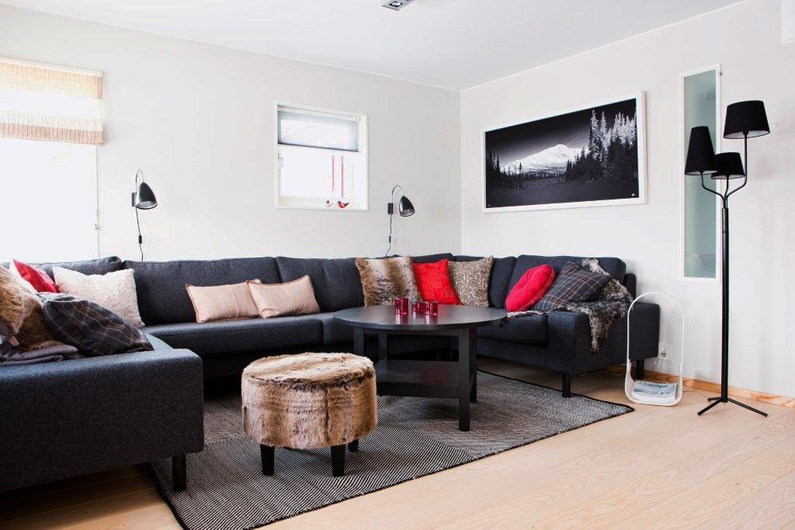En moderne stue med en mørk hjørnesofa, forskellige puder, et rundt sofabord og pelsbetrukne skamler. Der er to små vinduer, et sort-hvidt landskabsfoto og en gulvlampe med flere skærme.