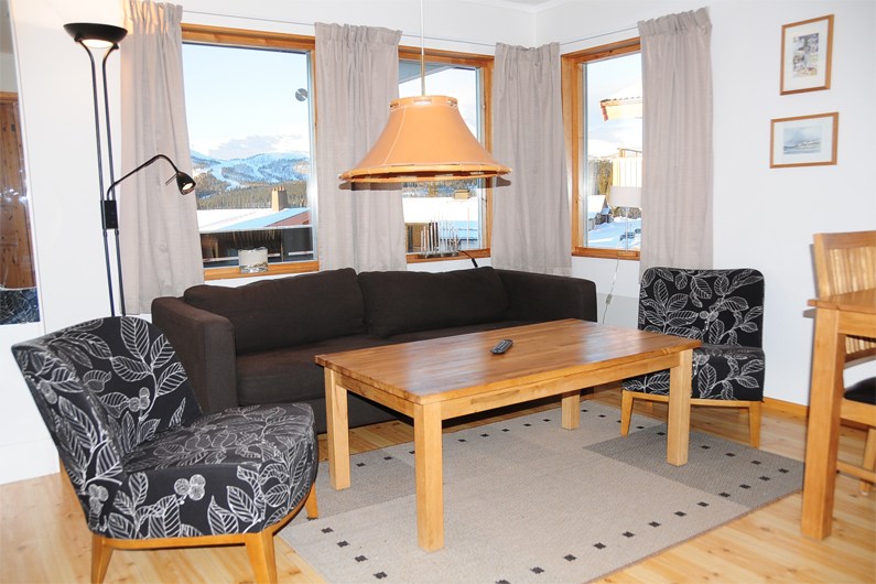 En hyggelig stue med en mørk sofa, to mønstrede lænestole, et sofabord i træ og et beige tæppe. Store vinduer med lyse gardiner afslører sneklædte bjerge og bygninger udenfor. En pendellampe hænger over bordet.