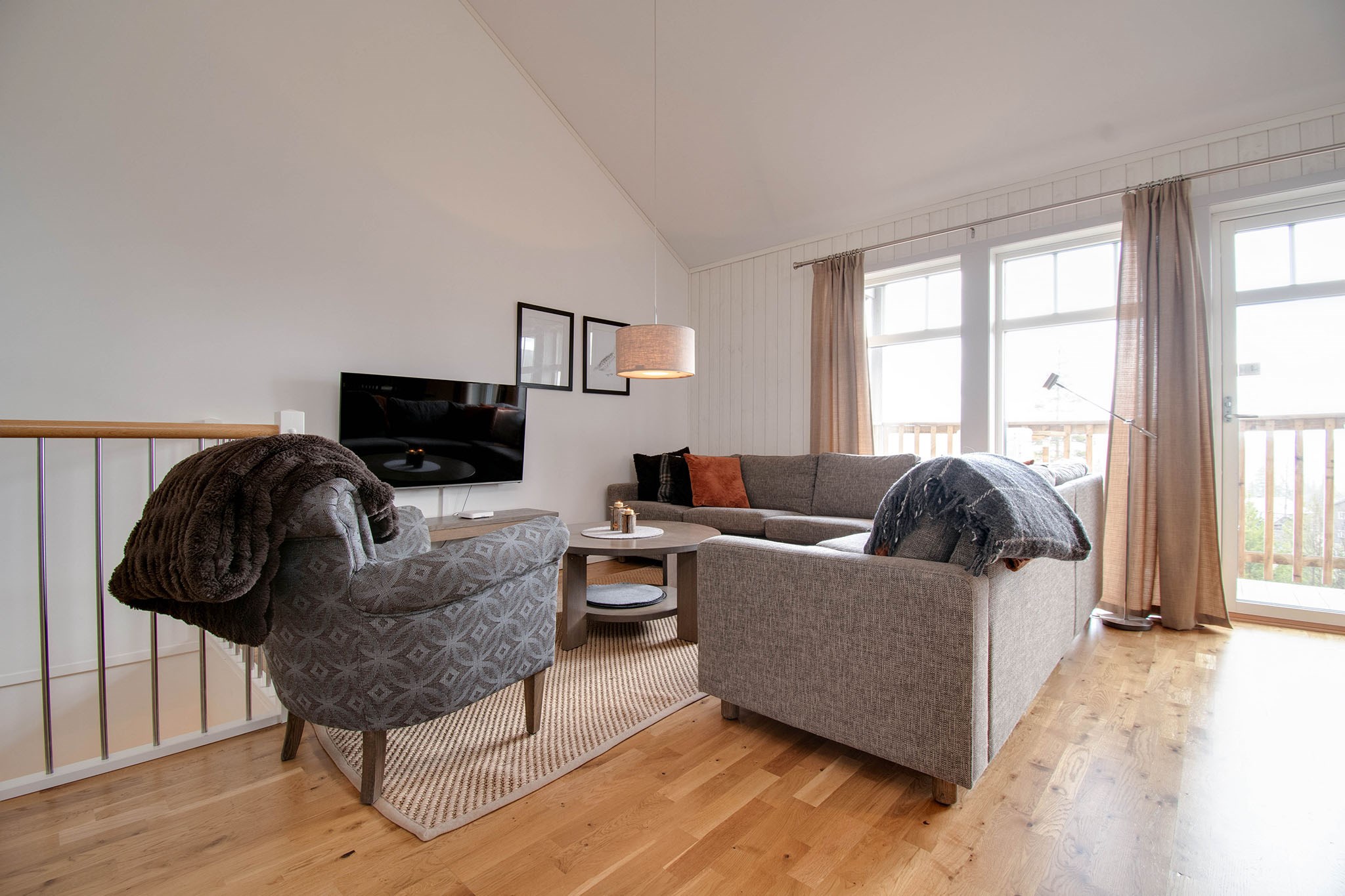 En hyggelig stue med en grå hjørnesofa, en mønstret lænestol, et sofabord i træ, et TV på væggen og store vinduer med beige gardiner, der lukker naturligt lys ind. Gulvet er af træ, og indretningen er moderne og minimalistisk.