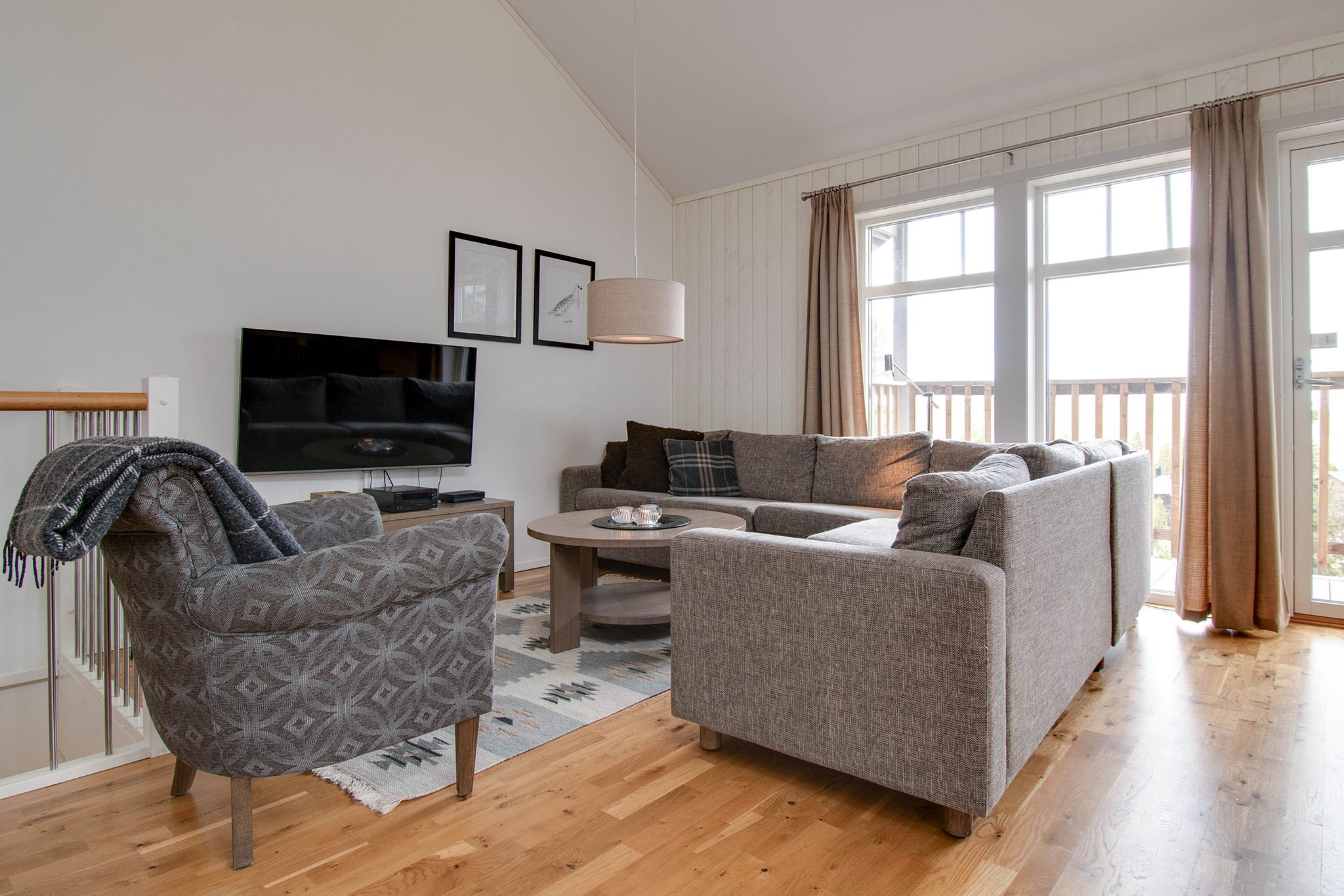 En moderne stue med en grå sofa og en mønstret lænestol, et rundt sofabord, et vægmonteret tv, indrammet kunst og store vinduer med beige gardiner, der lukker naturligt lys ind.