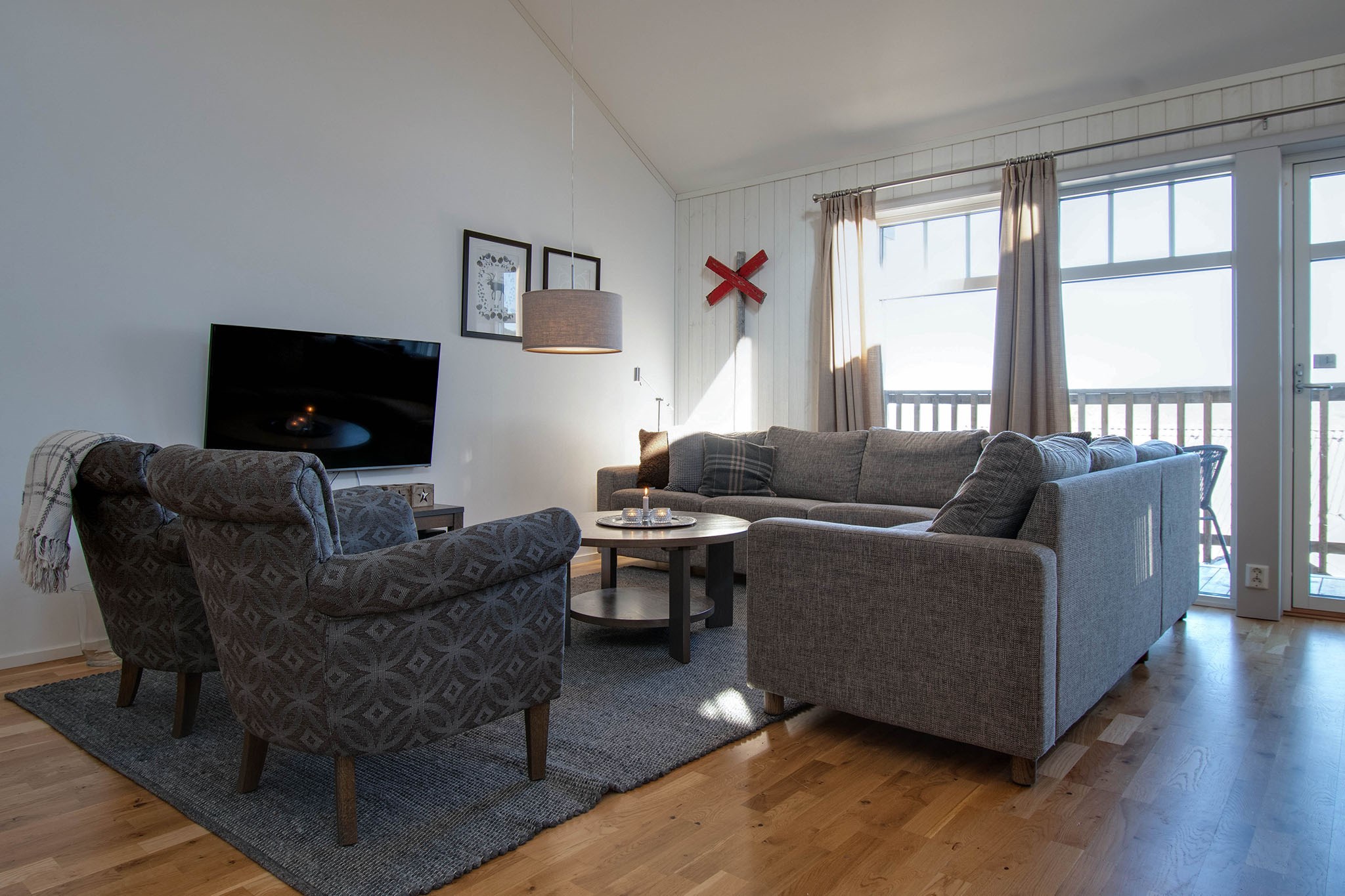 En moderne stue med en grå hjørnesofa, to mønstrede lænestole, et rundt sofabord, et TV på et stativ, store vinduer med gardiner og trægulv. Sollys filtreres ind og skaber en lys atmosfære.