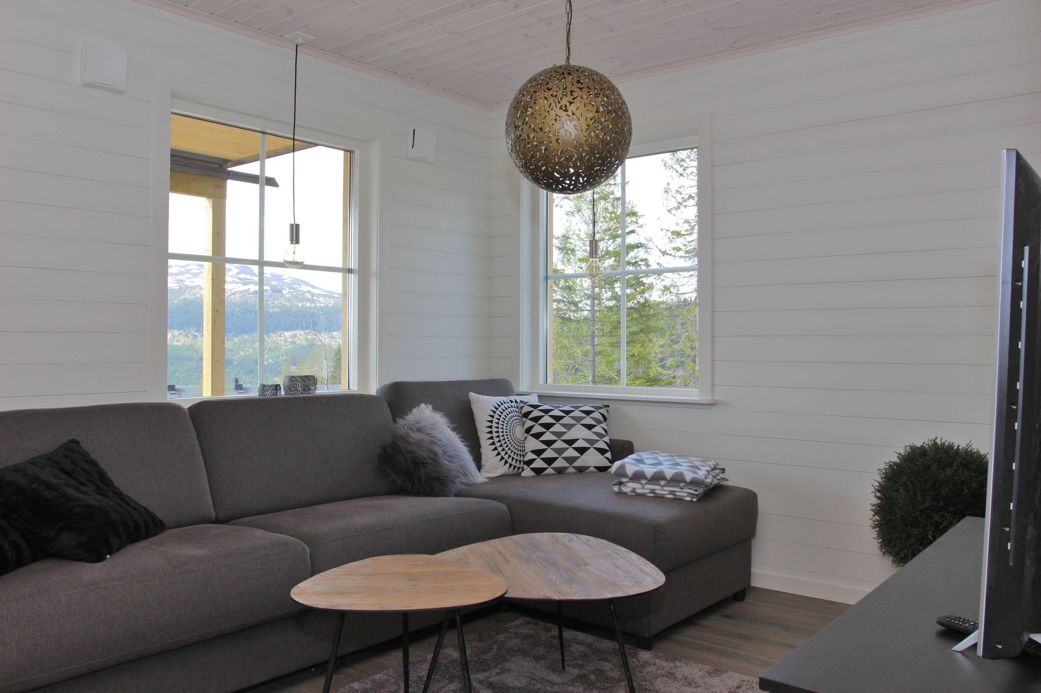 En moderne stue med en grå hjørnesofa, geometriske og pelspuder, to runde sofaborde i træ, en stor dekorativ pendellampe og vinduer med en smuk udsigt over bjergene.