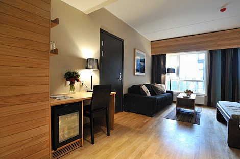 Moderne hotelværelse med lyse trægulve, et lille skrivebord med stol og lampe, et minikøleskab, en mørk sofa, et tæppe, en seng og et stort vindue, der lukker naturligt lys ind. Indretningen er enkel og moderne.